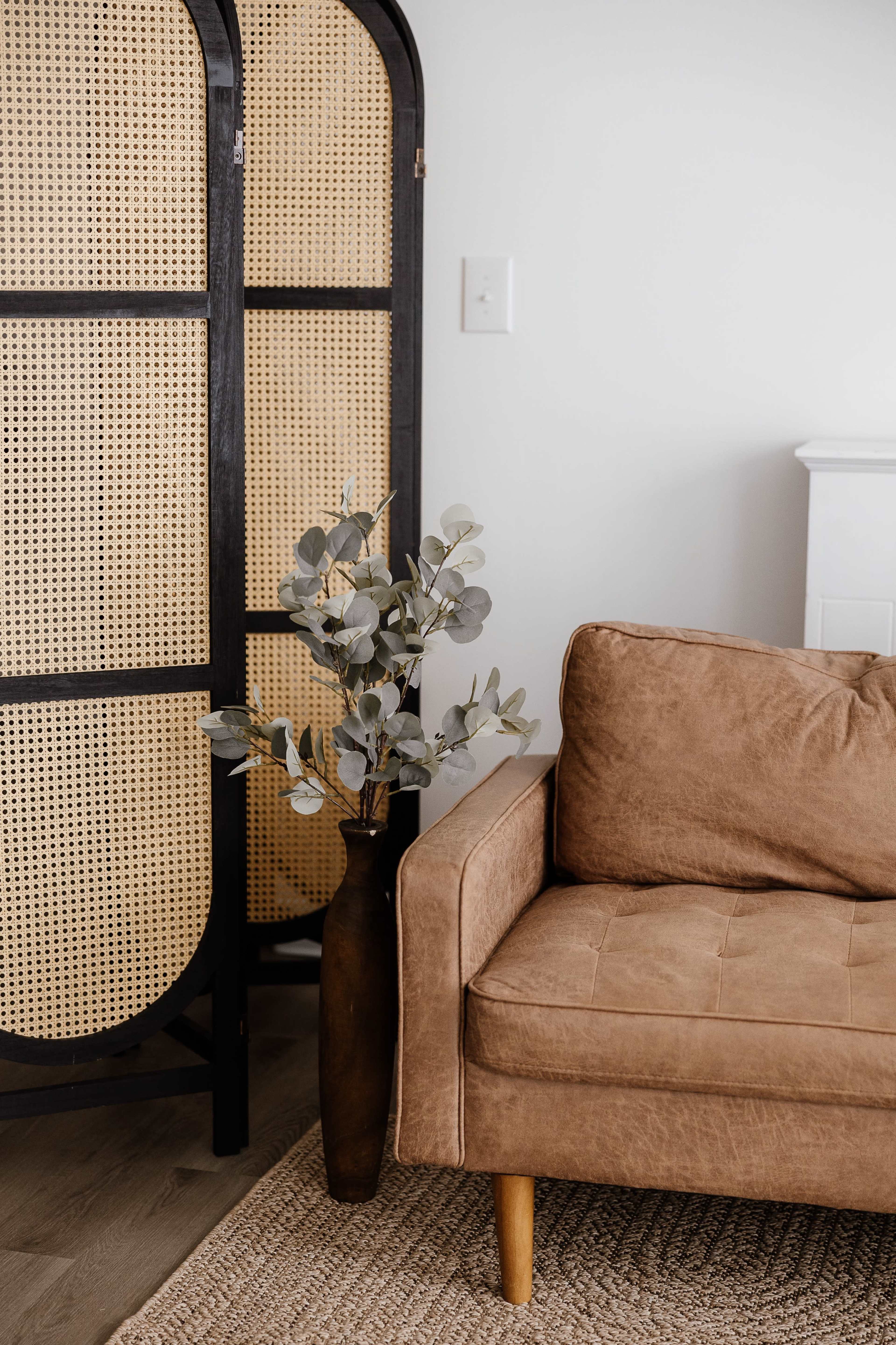 The image shows a brown couch next to a tall, dark vase containing eucalyptus branches, alongside a woven room divider.