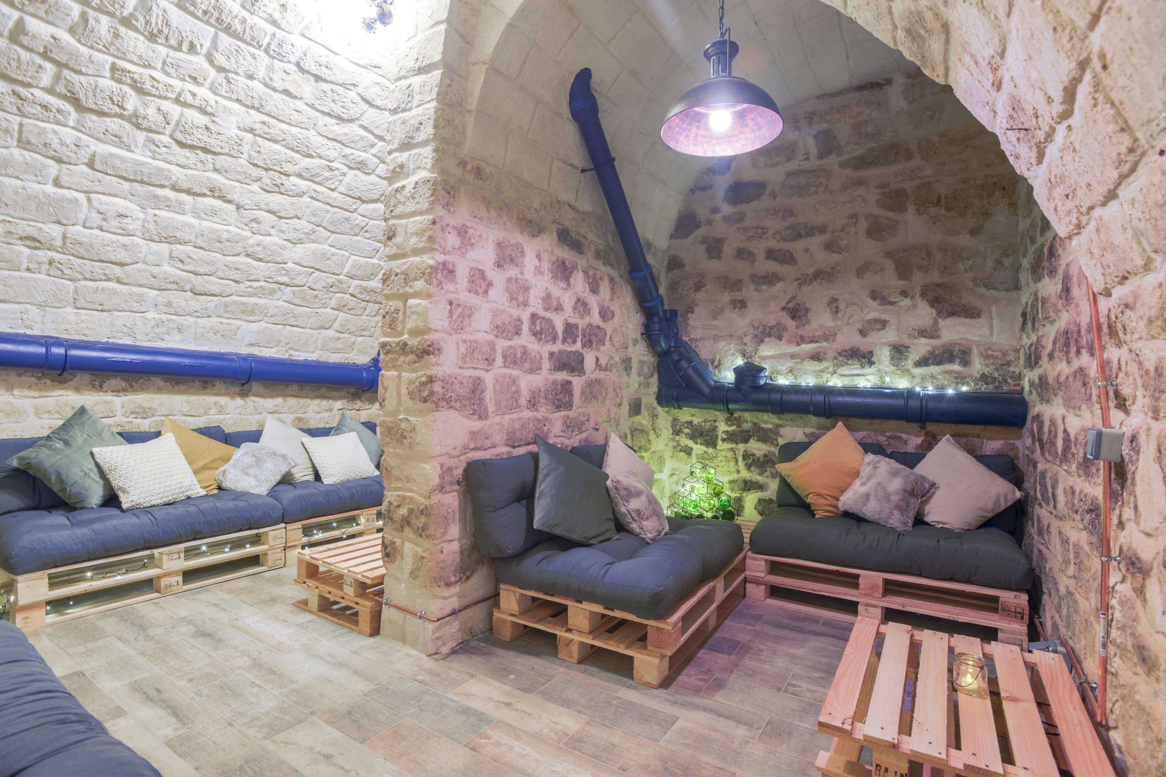 A cozy, underground seating area with stone walls and wooden pallet furniture arranged around soft cushions.