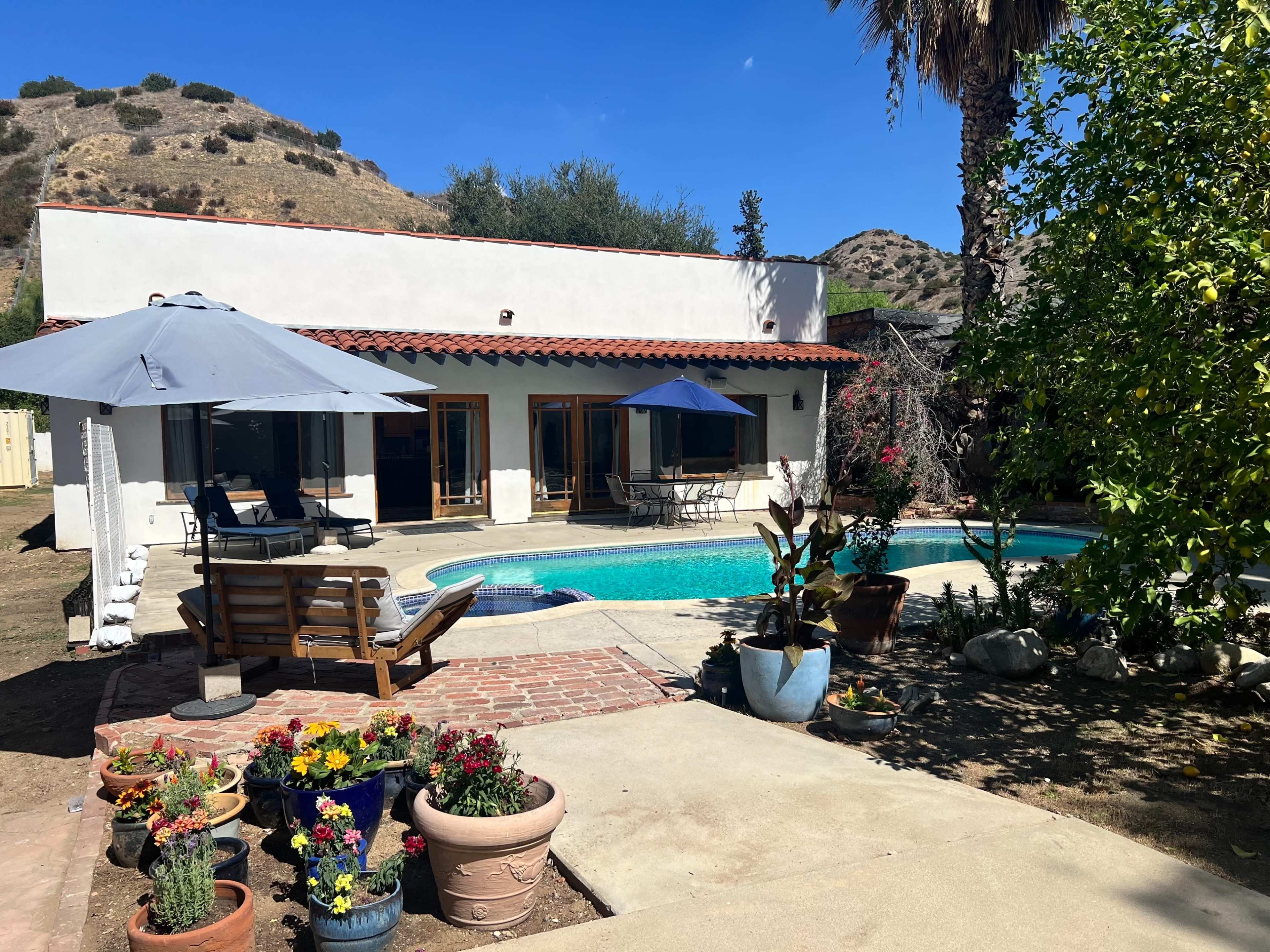 A white stucco house with a red tile roof and a pool is surrounded by potted plants and patio furniture in a yard with rolling hills in the background.
