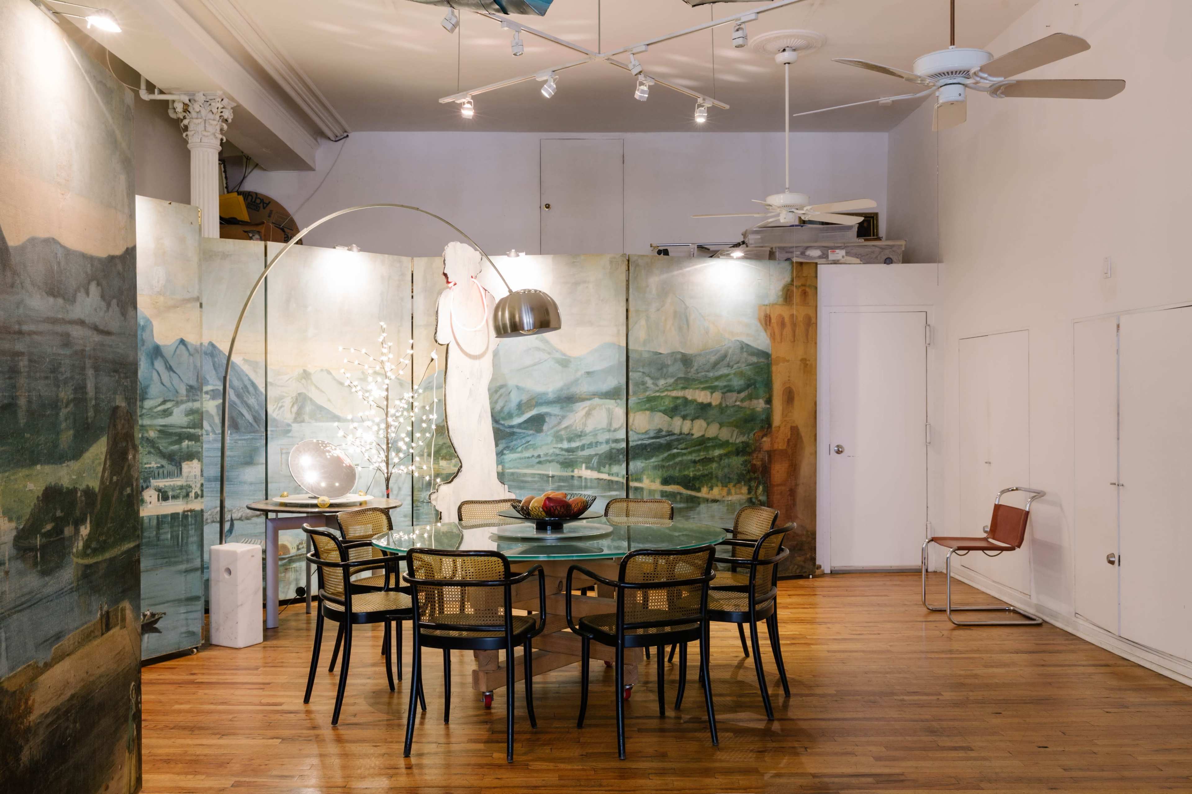 A spacious interior features a round glass dining table surrounded by chairs, with large mural artworks on the walls and a decorative arc lamp overhead.