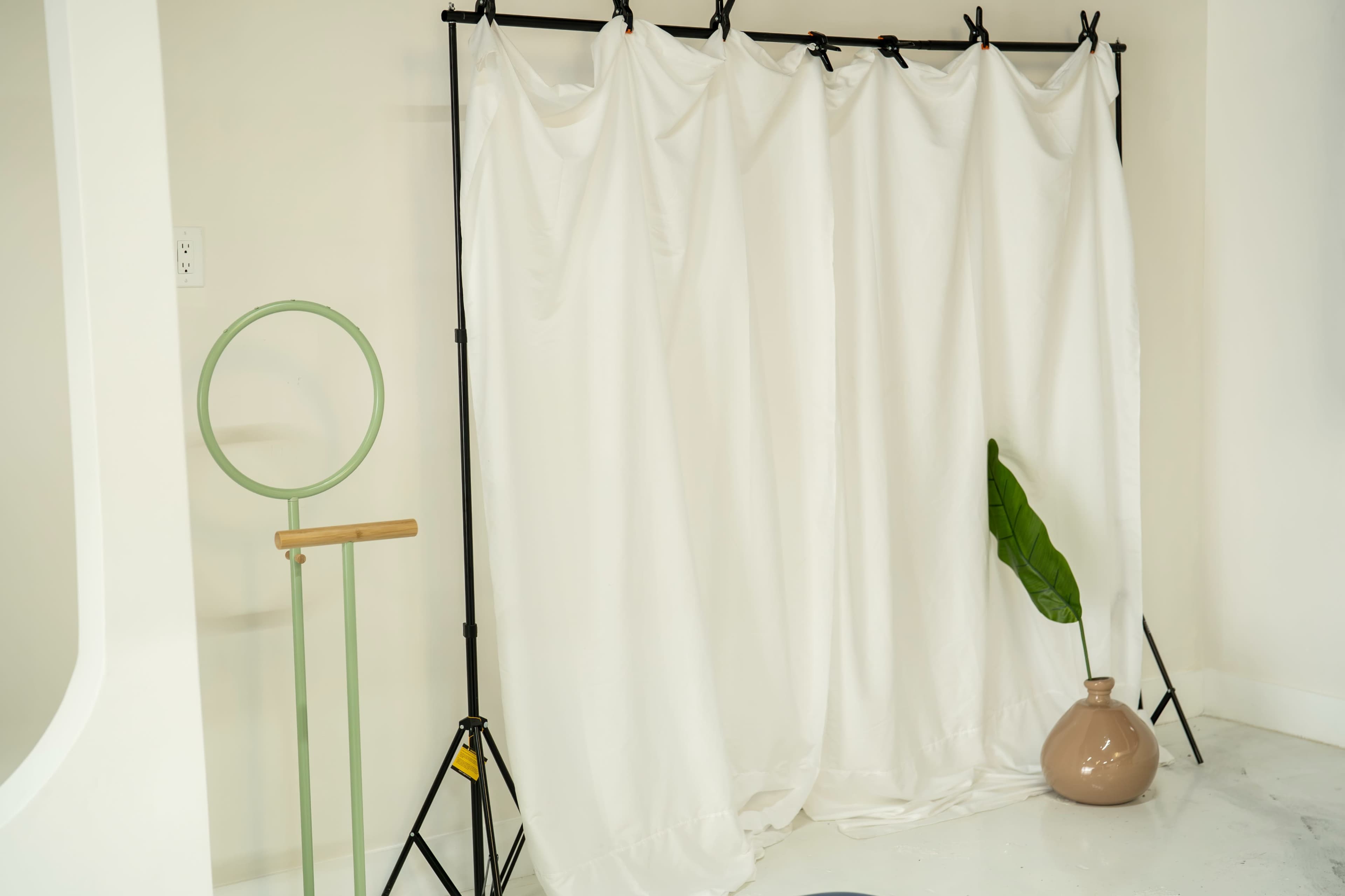 A white fabric backdrop is draped on a black frame, accompanied by a green plant and a brown pottery vase on the floor.