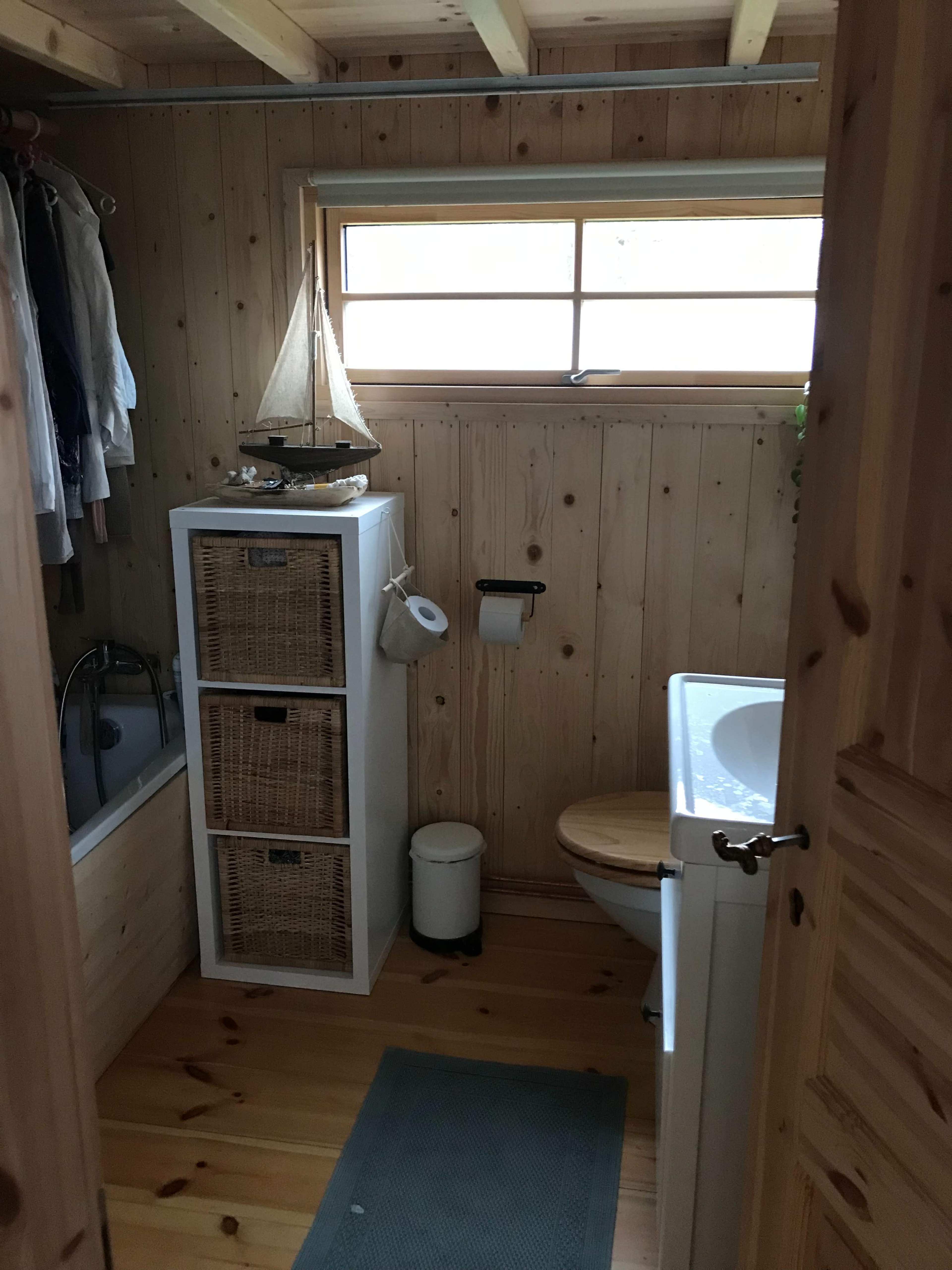 The image shows a small bathroom with wooden walls, a shower area, a sink, a cabinet for storage, and a sailboat decoration on top of the cabinet.
