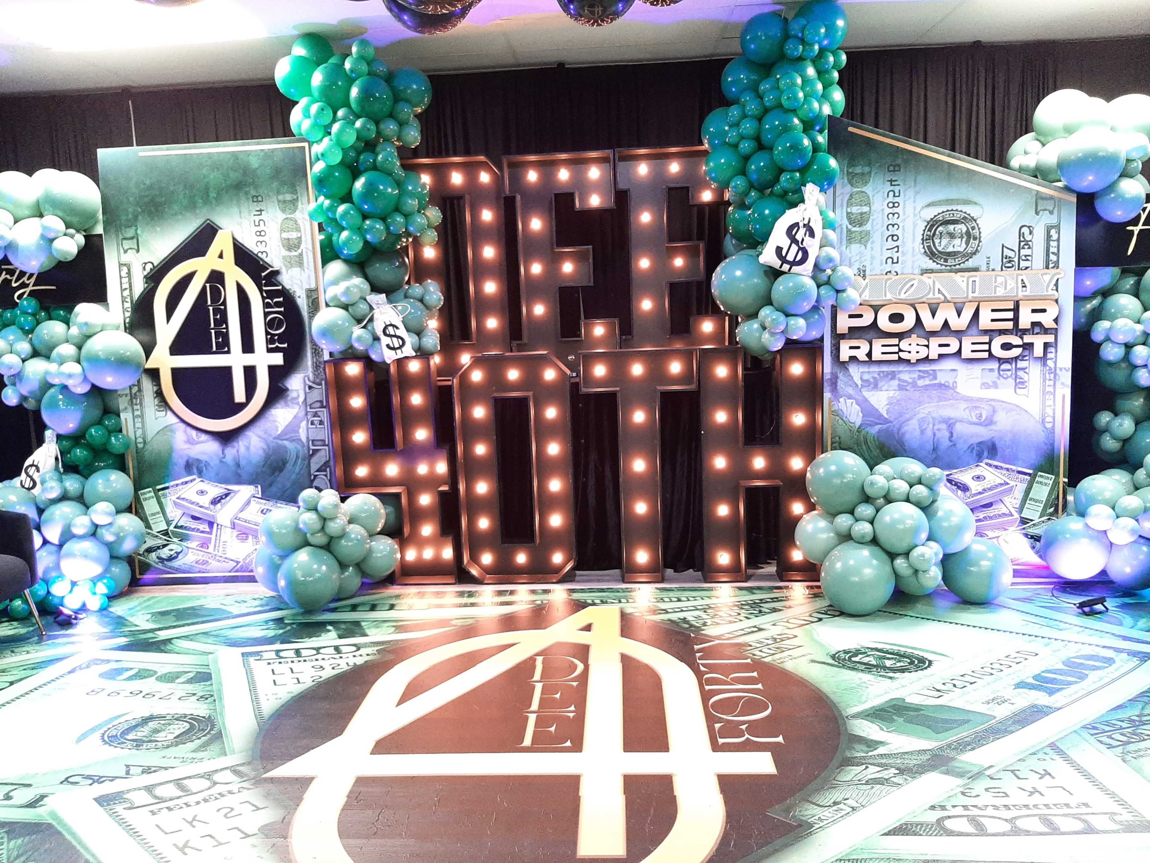 A party backdrop with large illuminated letters spelling "DEE 40TH," surrounded by green and teal balloons, and dollar-themed decorations on the floor.