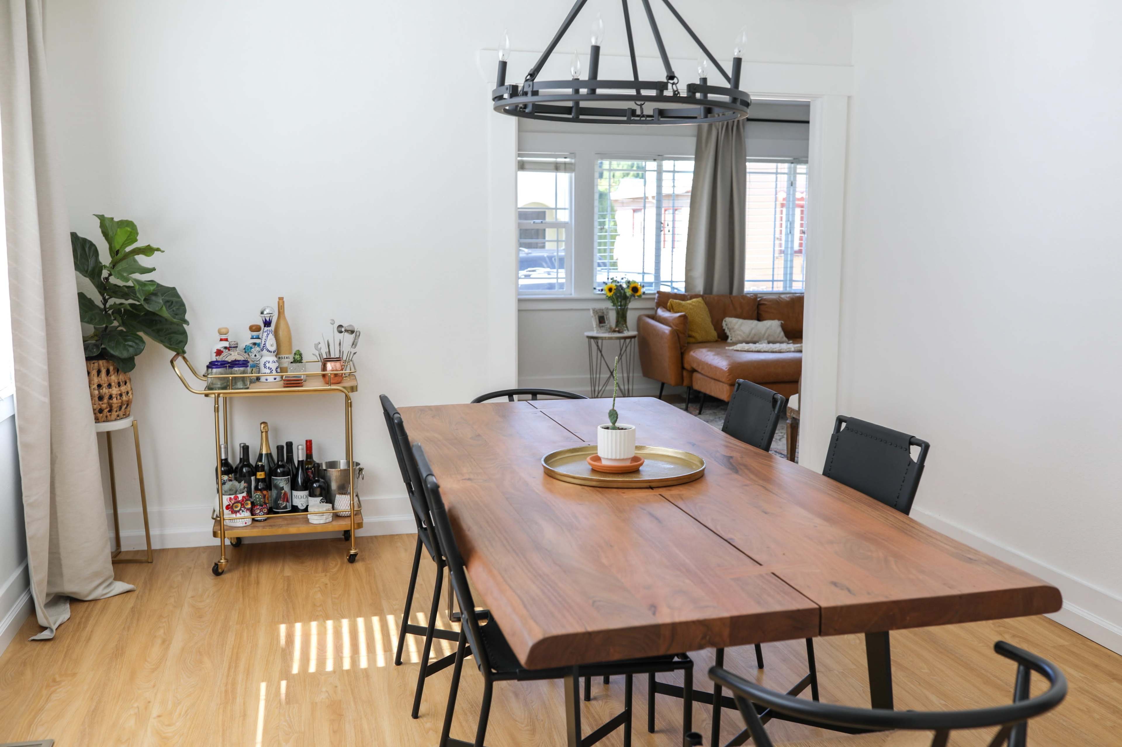 A wooden dining table with black chairs is positioned in a bright room featuring a plant and a bar cart with various beverages.