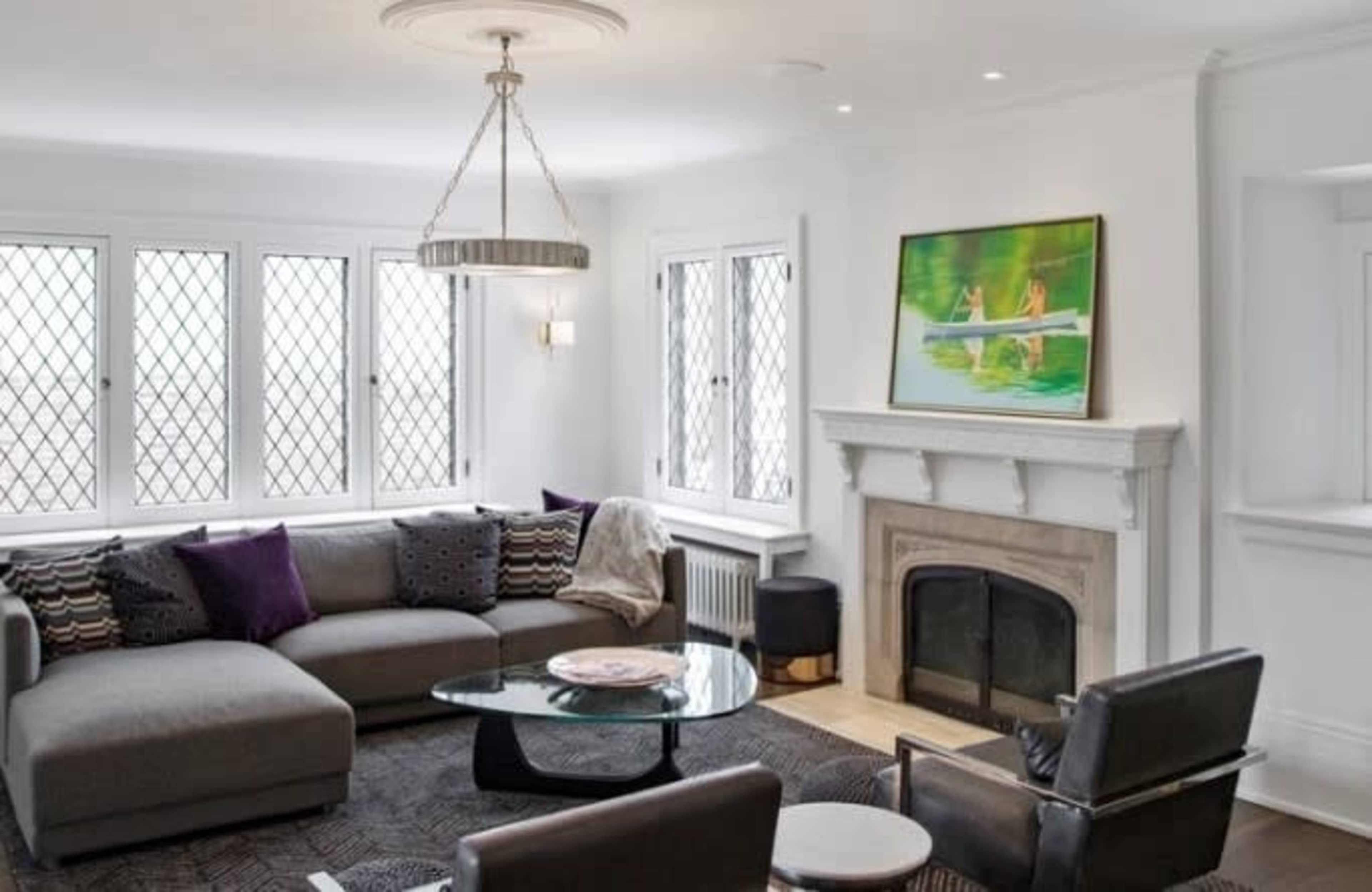 A modern living room features a gray sectional sofa, a glass coffee table, and a fireplace beneath a decorative painting, all set against a backdrop of large diamond-patterned windows.