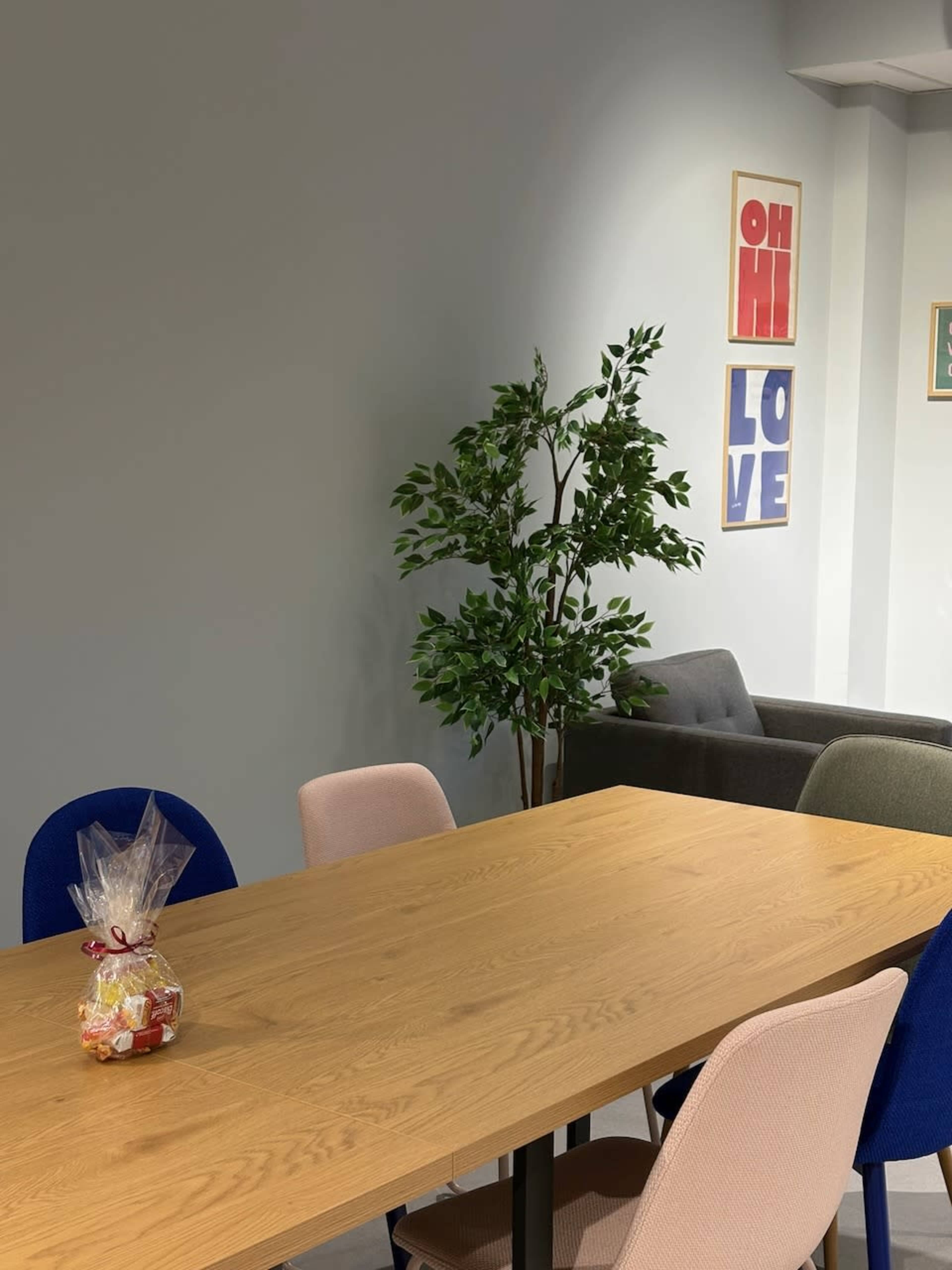 A wooden table with a colorful assortment of chairs is set in a modern meeting room with a potted plant and framed wall art in the background.