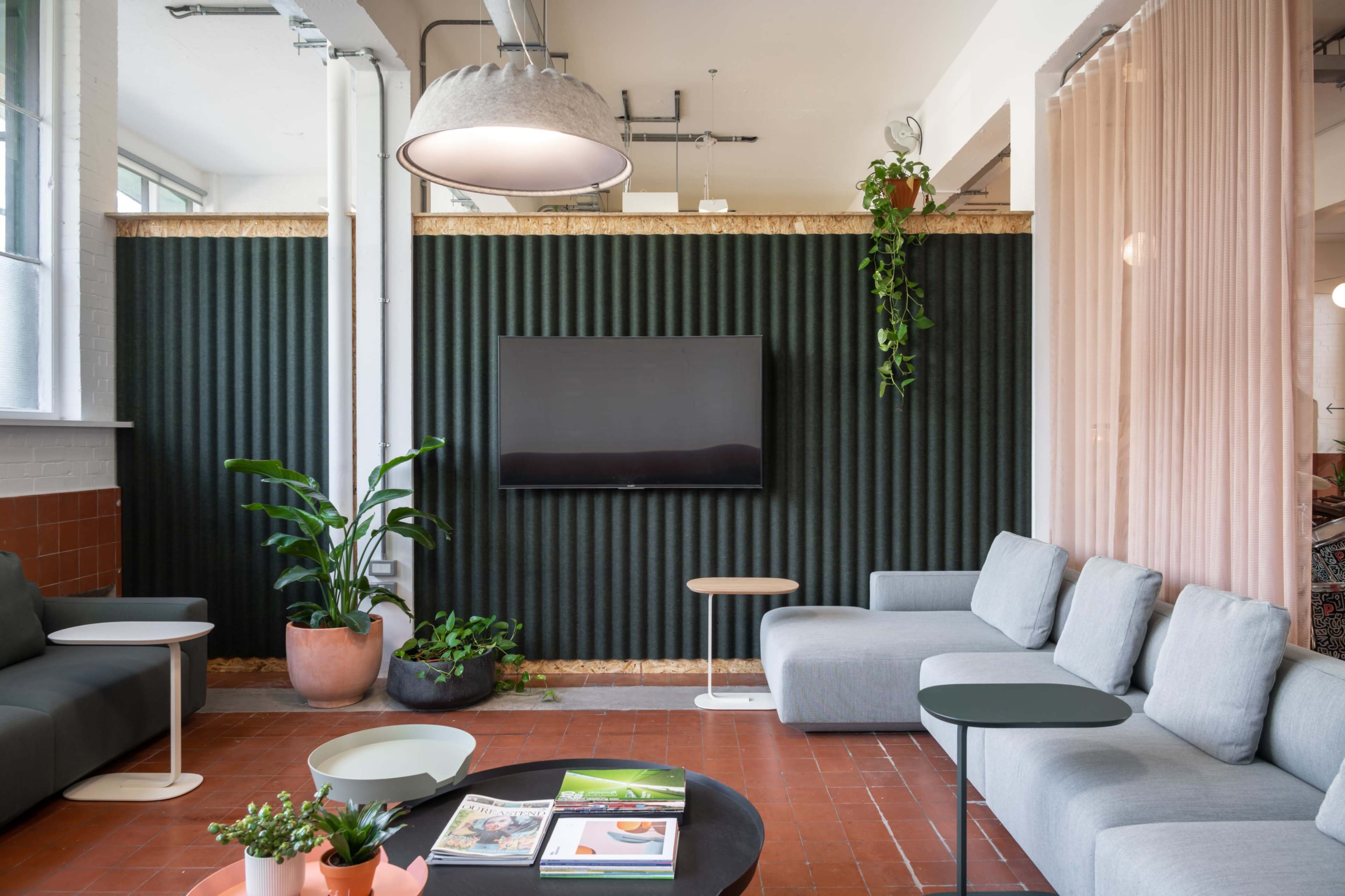 A modern lounge area with gray sofas, a wall-mounted television, and indoor plants against a textured green wall.