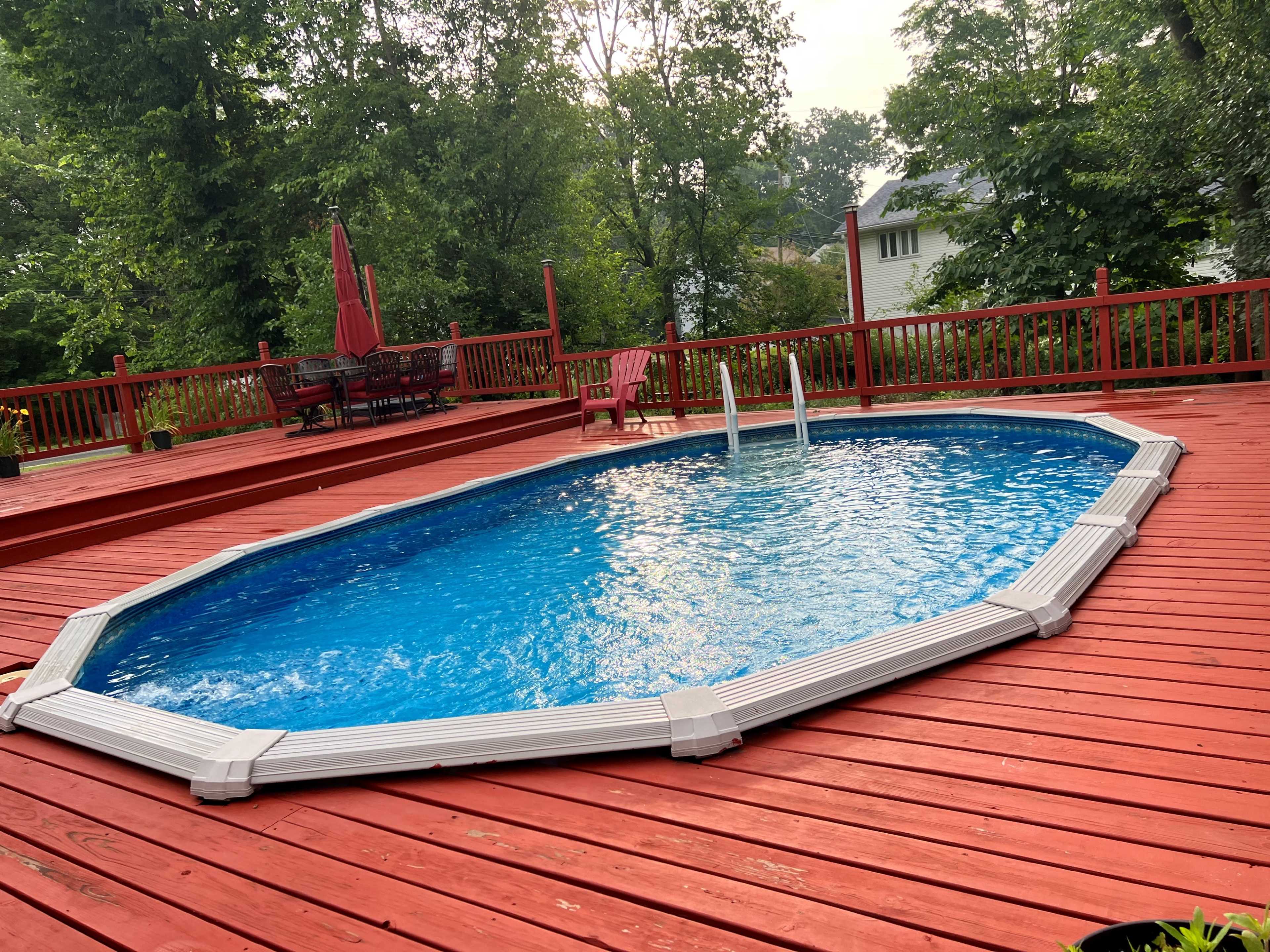 The image shows an oval above-ground pool with clear water surrounded by a red wooden deck and some patio furniture.