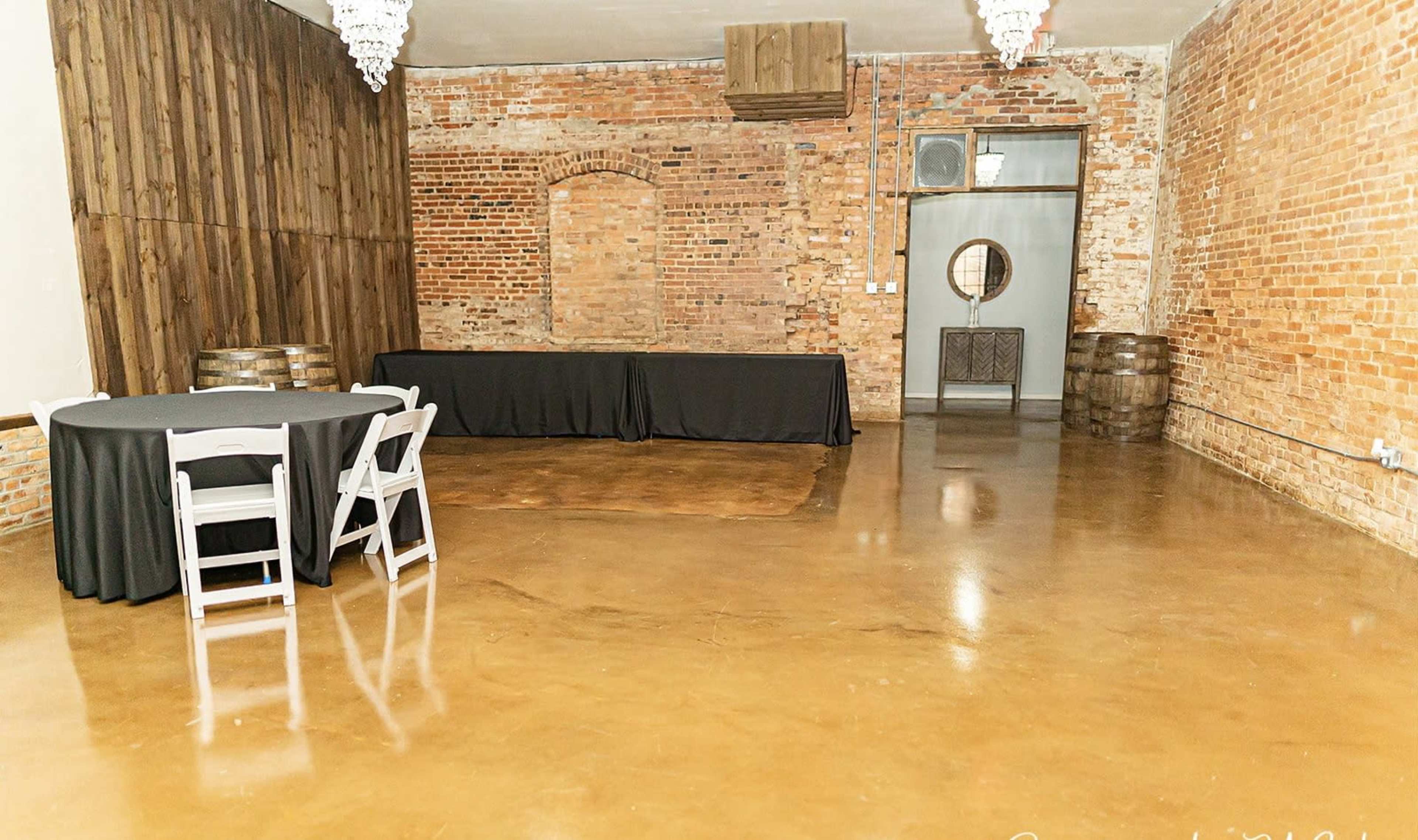 The image shows a spacious indoor venue featuring a polished concrete floor, exposed brick walls, and wooden accents, with a round table and a long black draped table against the wall.
