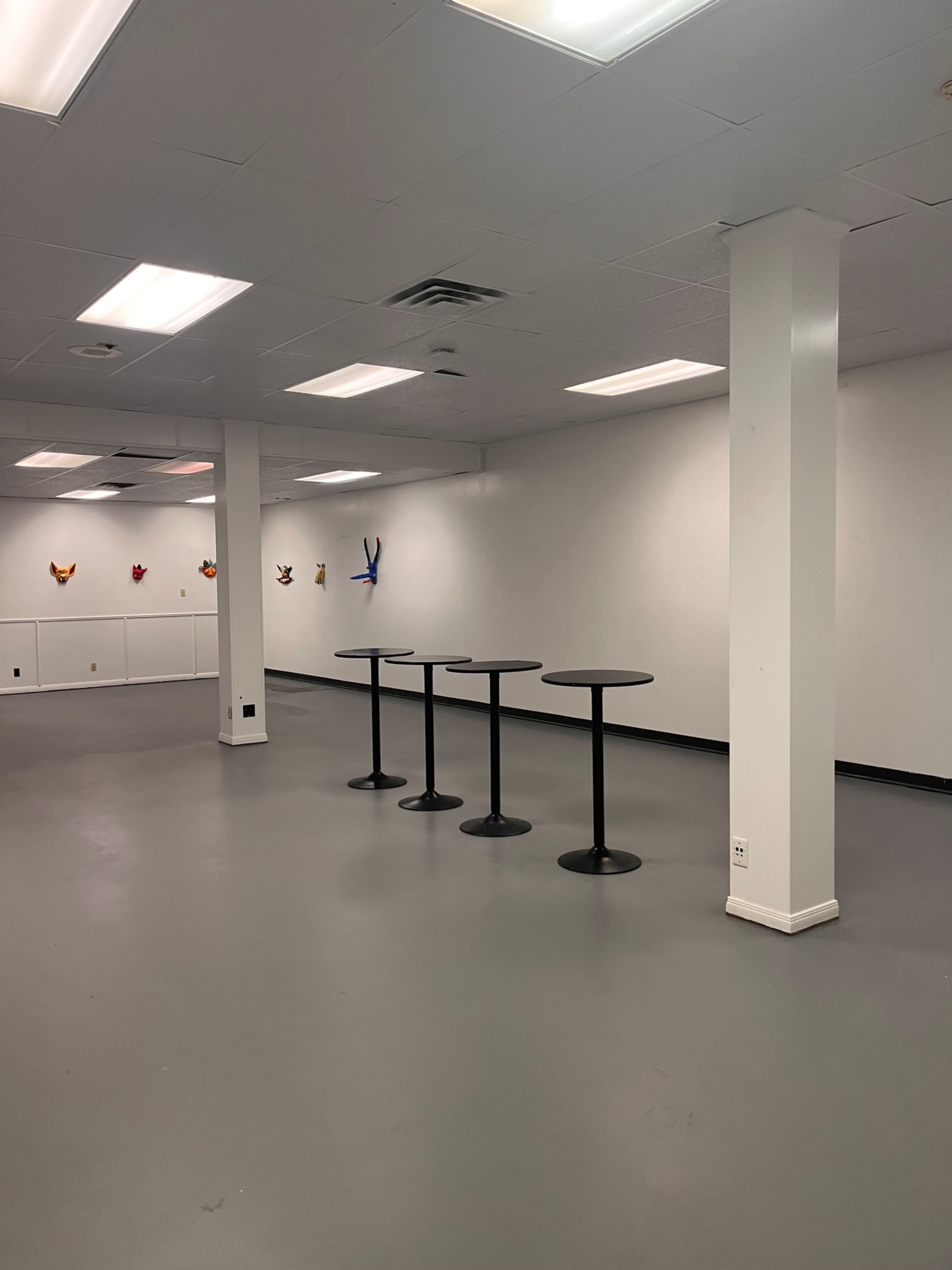 The image shows a spacious, well-lit room with gray flooring, featuring four round black tables and colorful wall decorations.