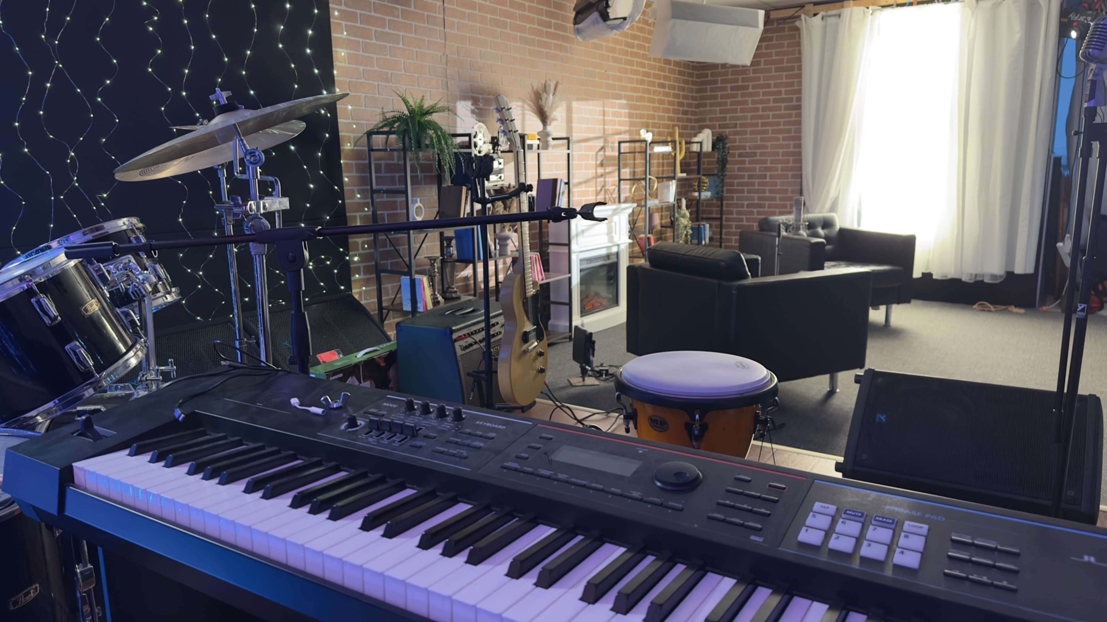 The image shows a music studio featuring a keyboard, drums, and various instruments, with a cozy seating area in the background and natural light coming through a window.