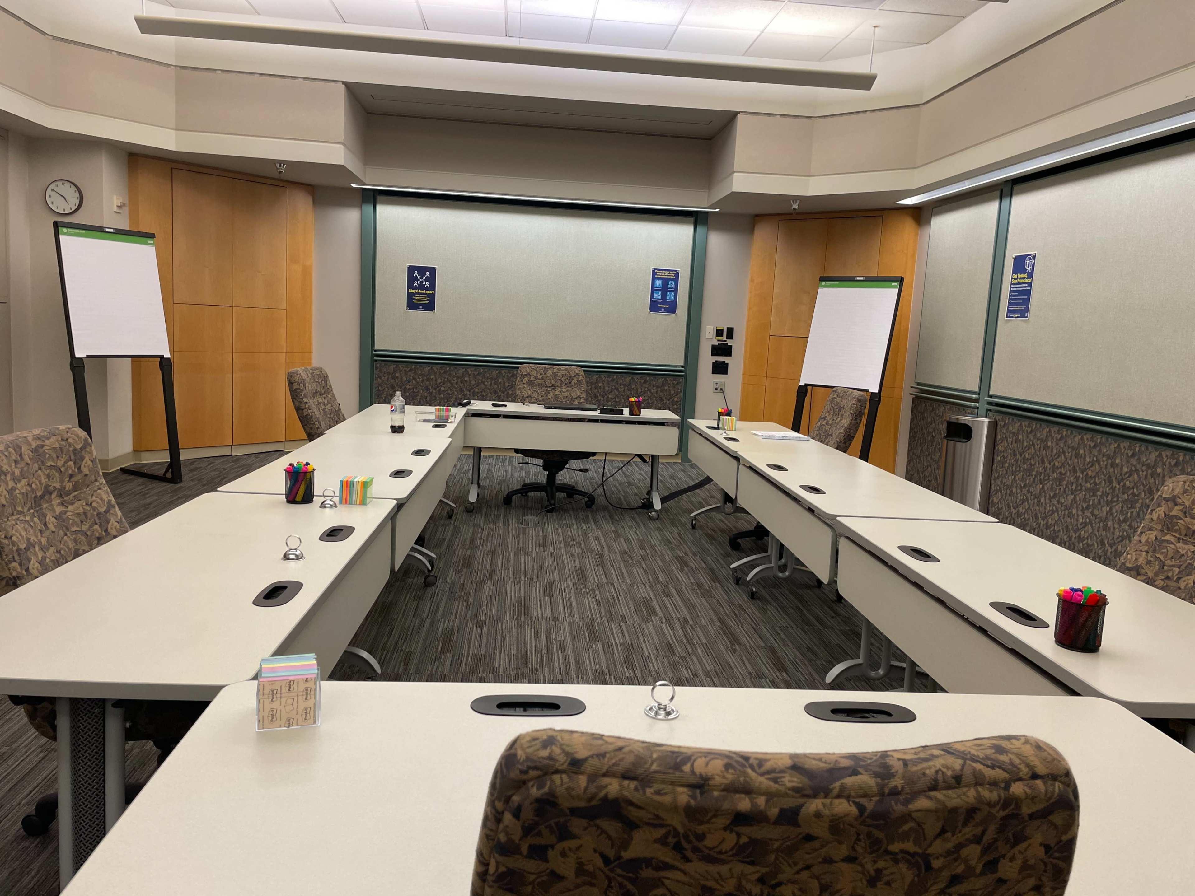A conference room features multiple tables arranged in a U-shape, with chairs, flip charts, and stationery neatly arranged.