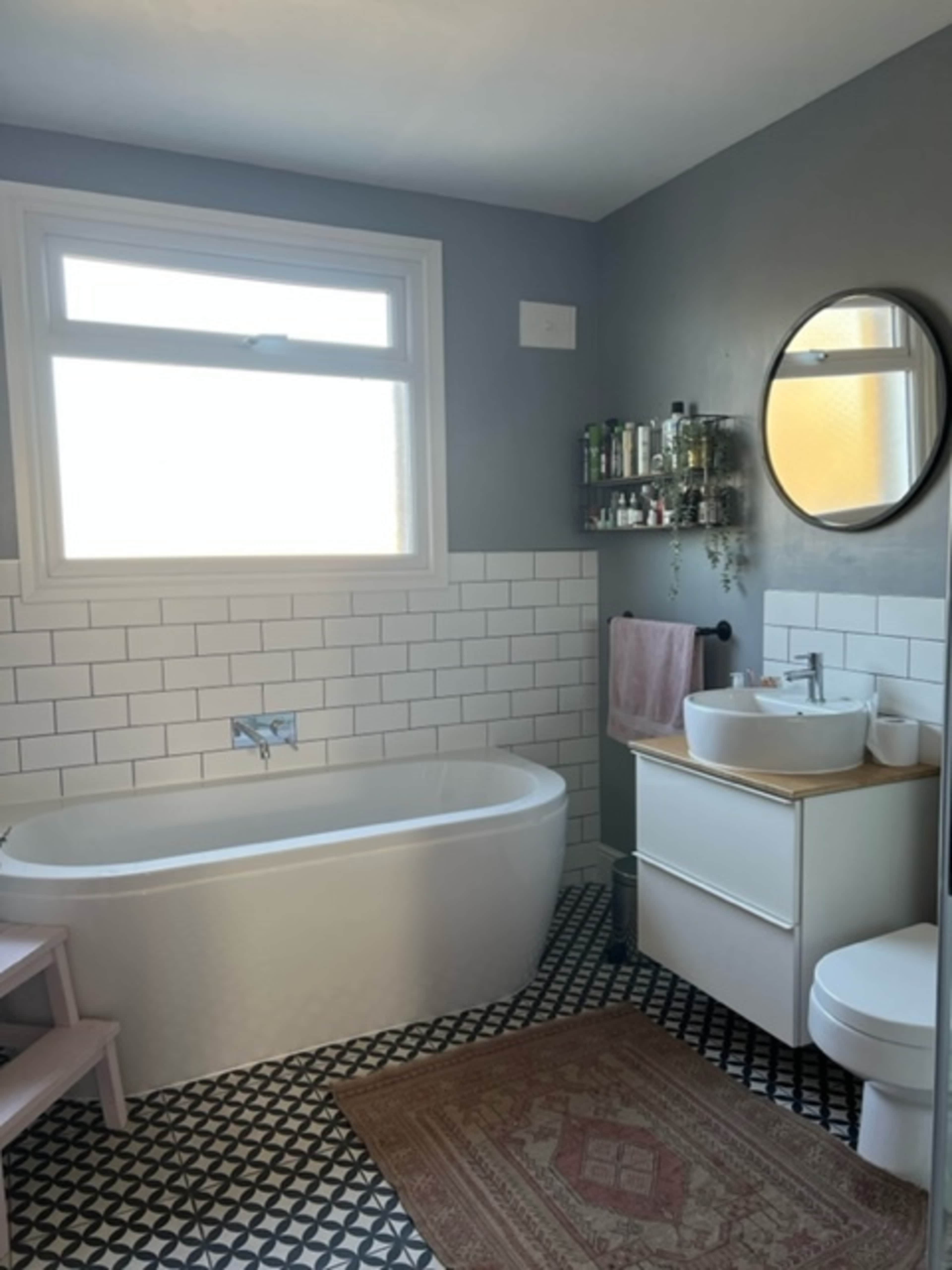 A modern bathroom featuring a freestanding bathtub, a circular mirror above a sink, a wall-mounted shelf with toiletries, and patterned floor tiles.