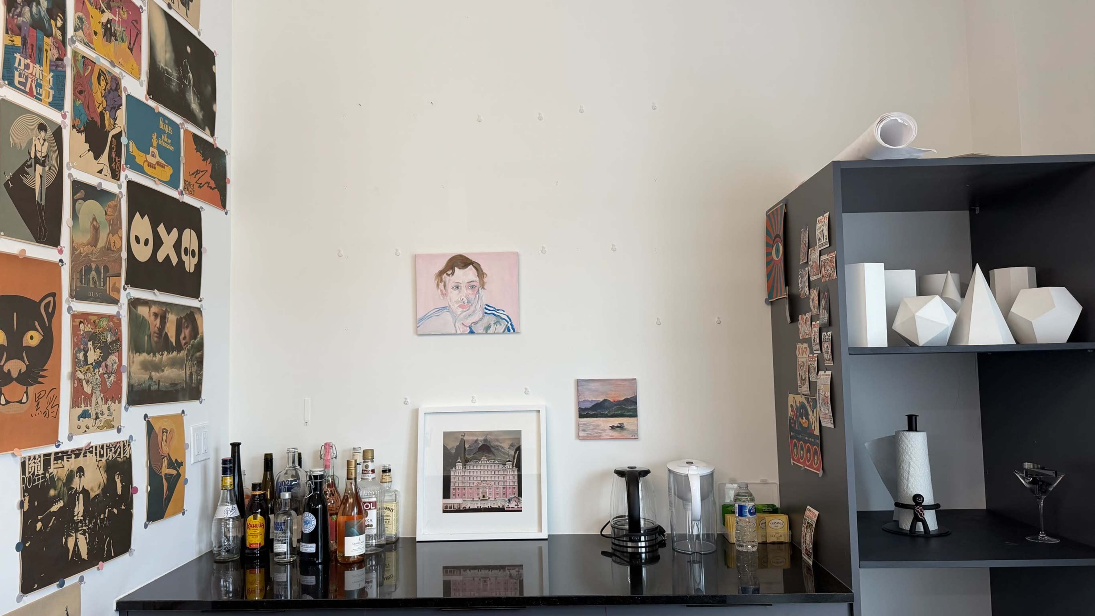 The image shows a corner of a room with a wall covered in artwork and a black countertop displaying various bottles and decorative items.