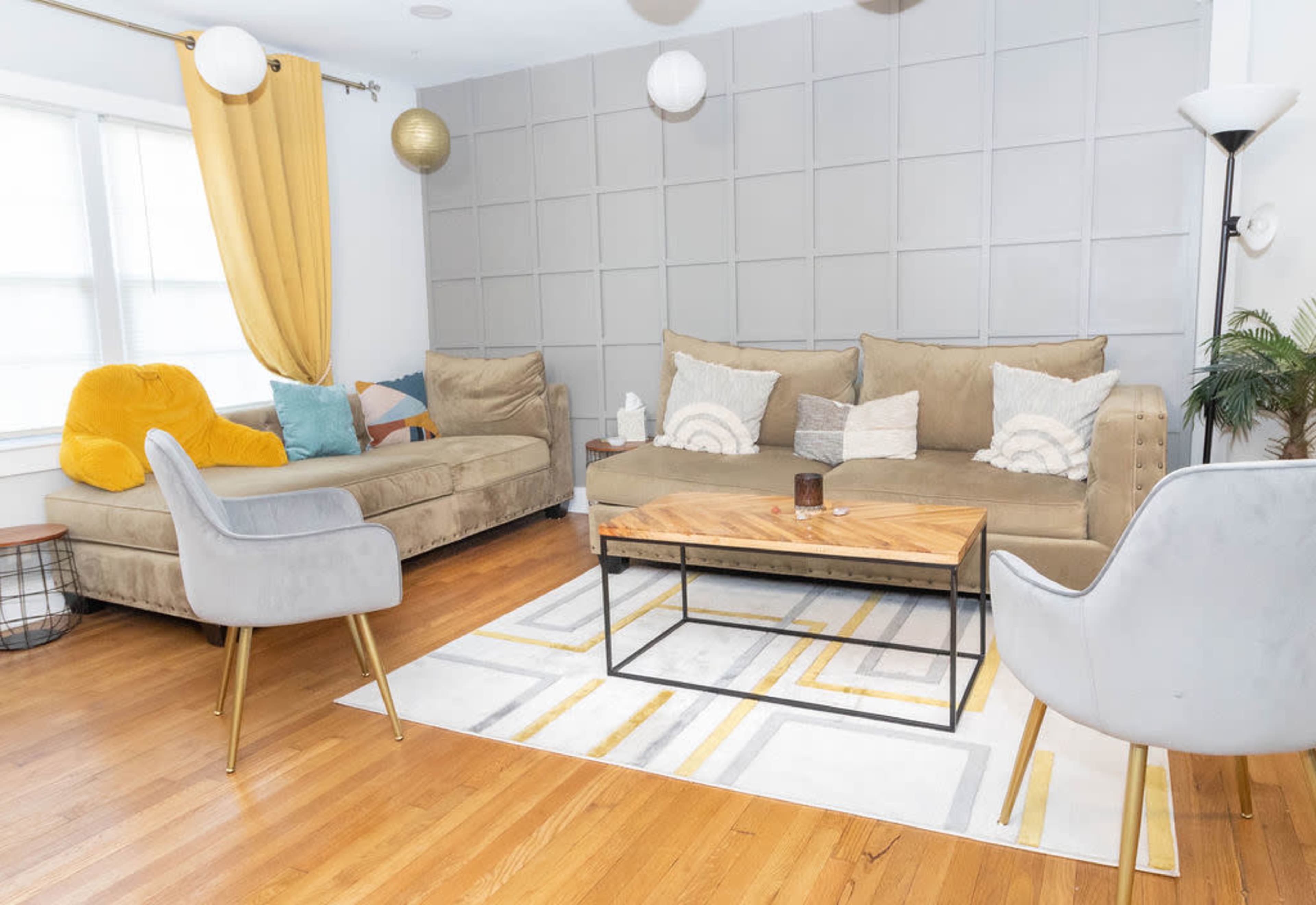 The image shows a living room featuring a sectional sofa, a wooden coffee table, and a gray rug with geometric patterns, complemented by large windows and decorative curtains.