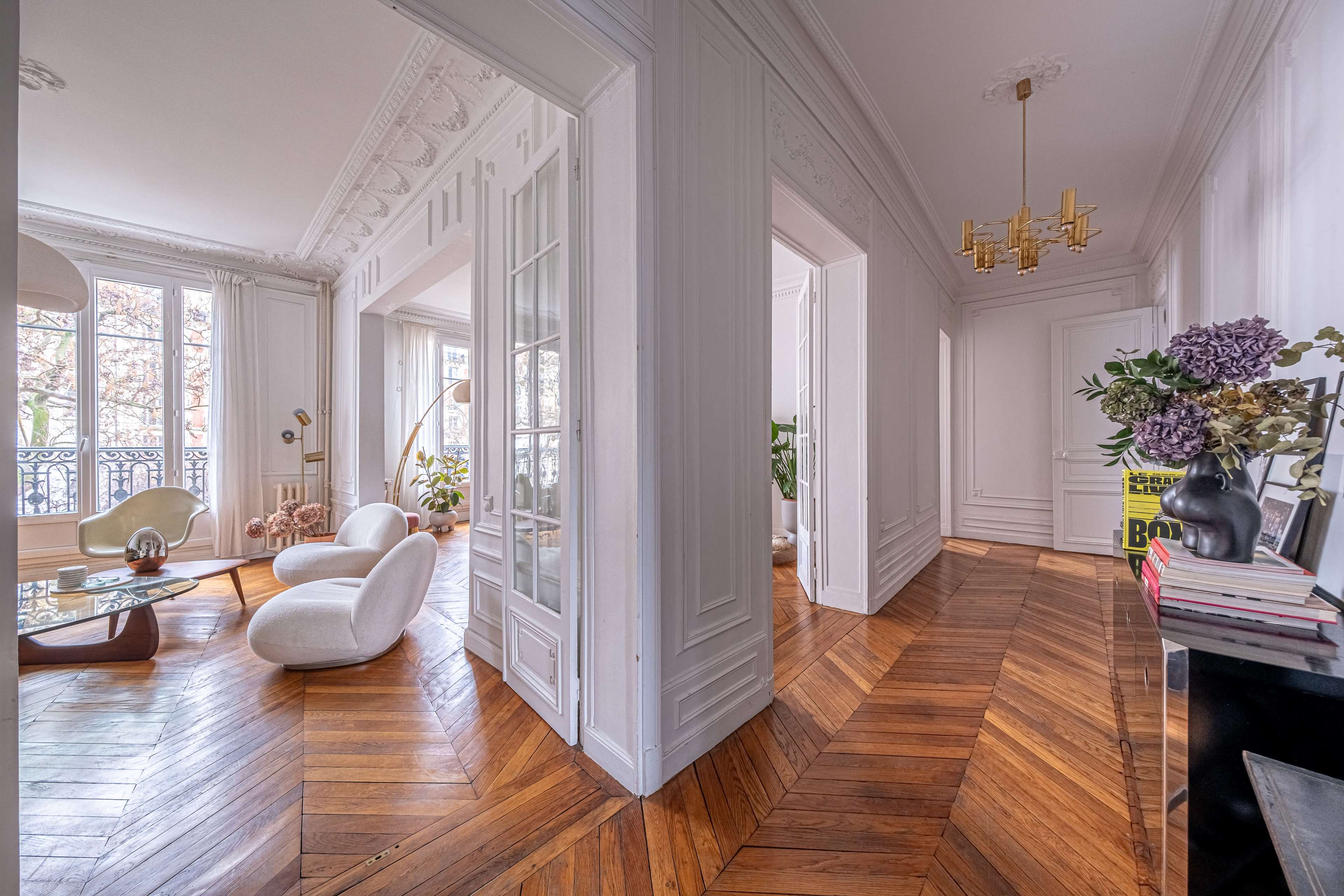 The image shows a spacious, elegantly designed interior with white walls, polished wooden flooring, and modern furniture arranged in a well-lit setting.