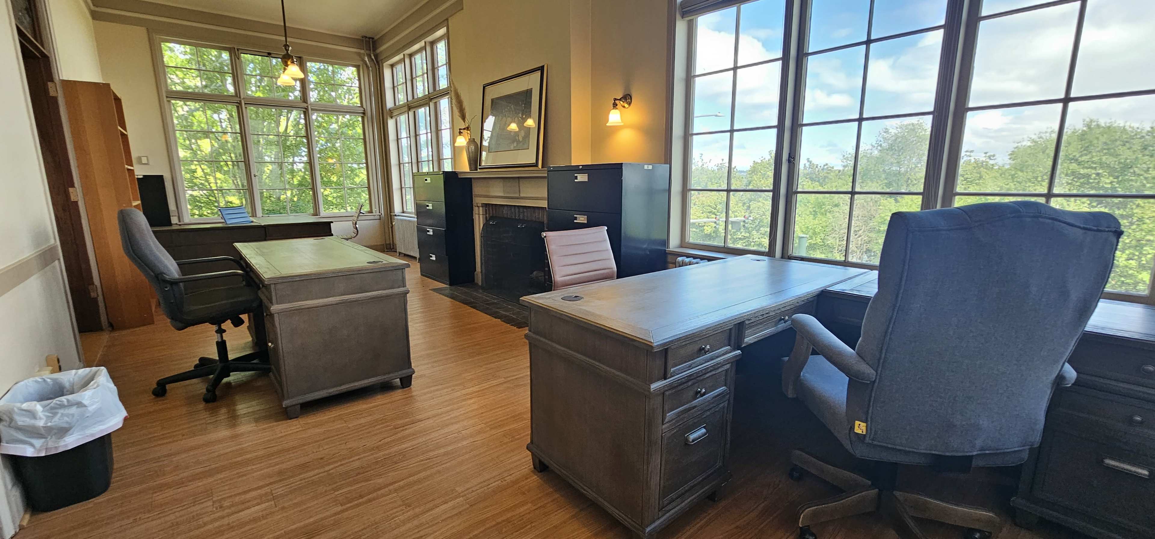 The image shows an office space with two desks, chairs, and large windows allowing natural light to enter, overlooking greenery outside.