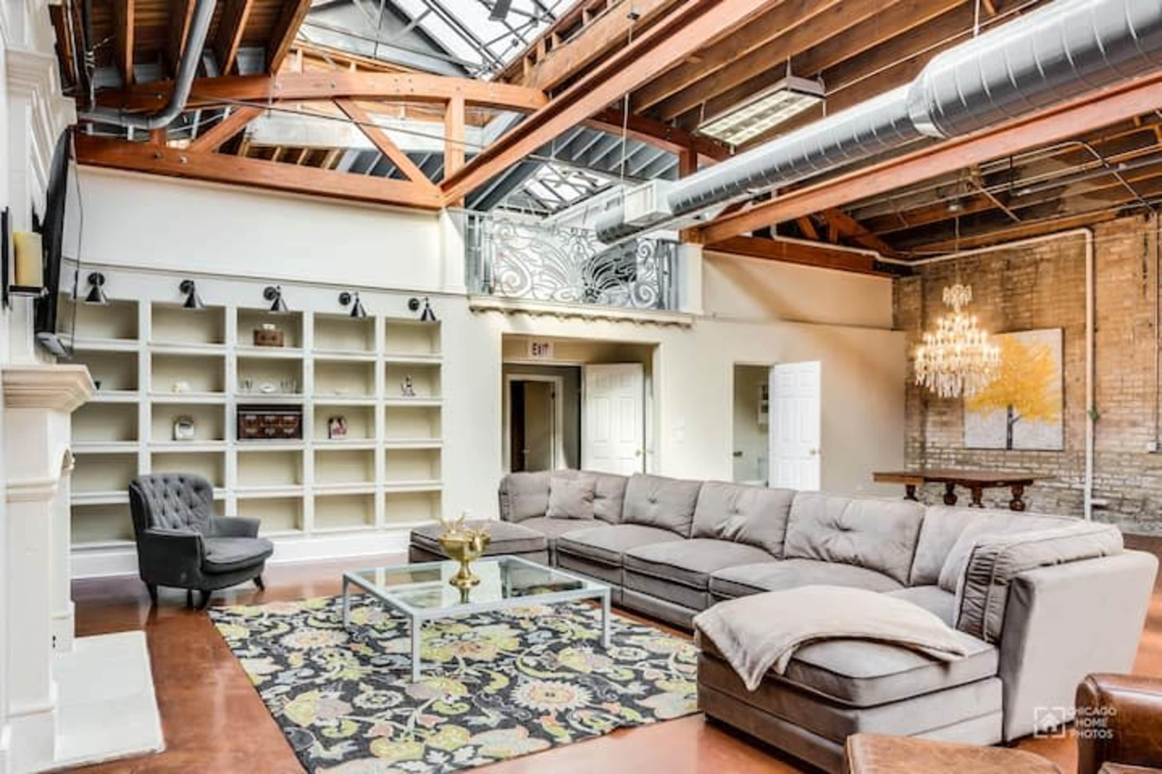 A spacious living area with a large sectional sofa, a coffee table, built-in shelves, and a chandelier, all beneath a skylight and wooden beams.