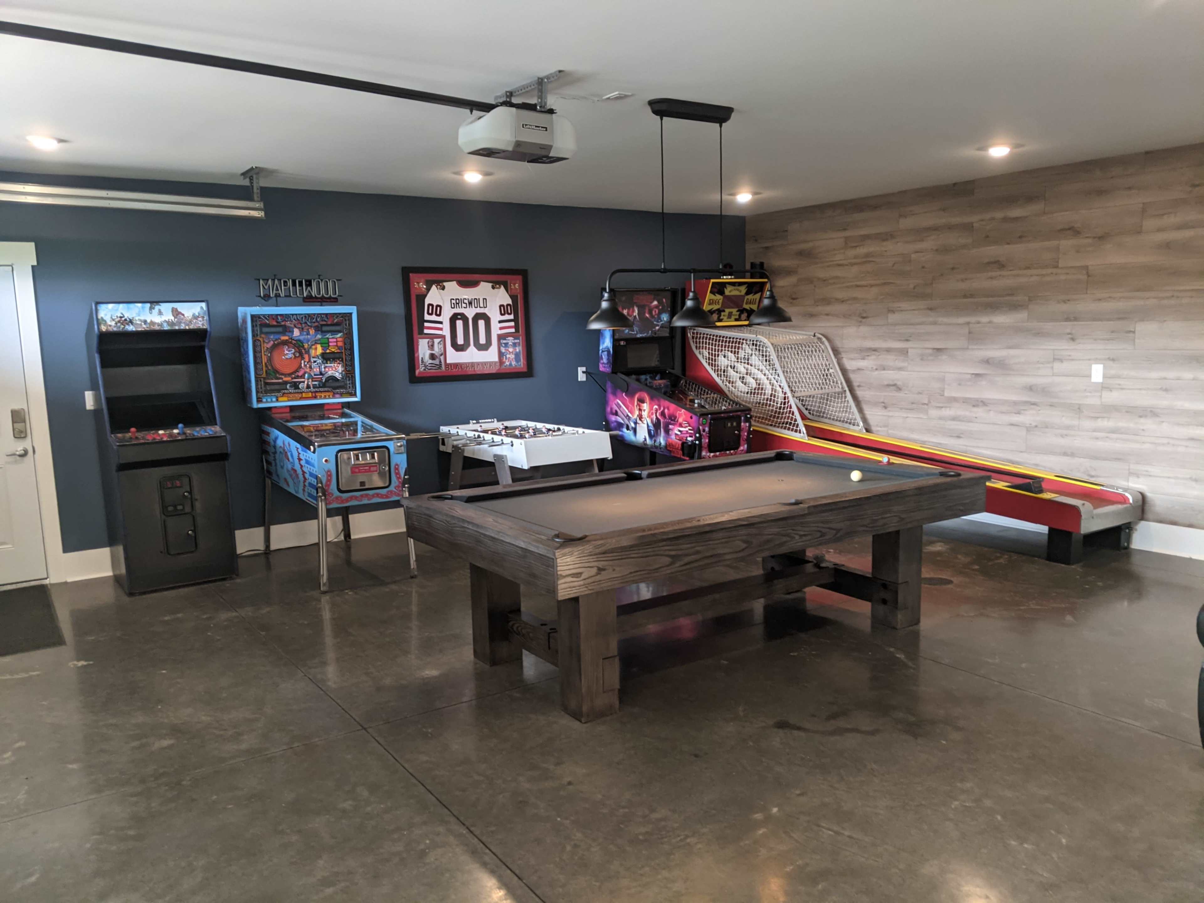 A game room features a billiards table, arcade machines, and a skee ball setup against a gray wall.