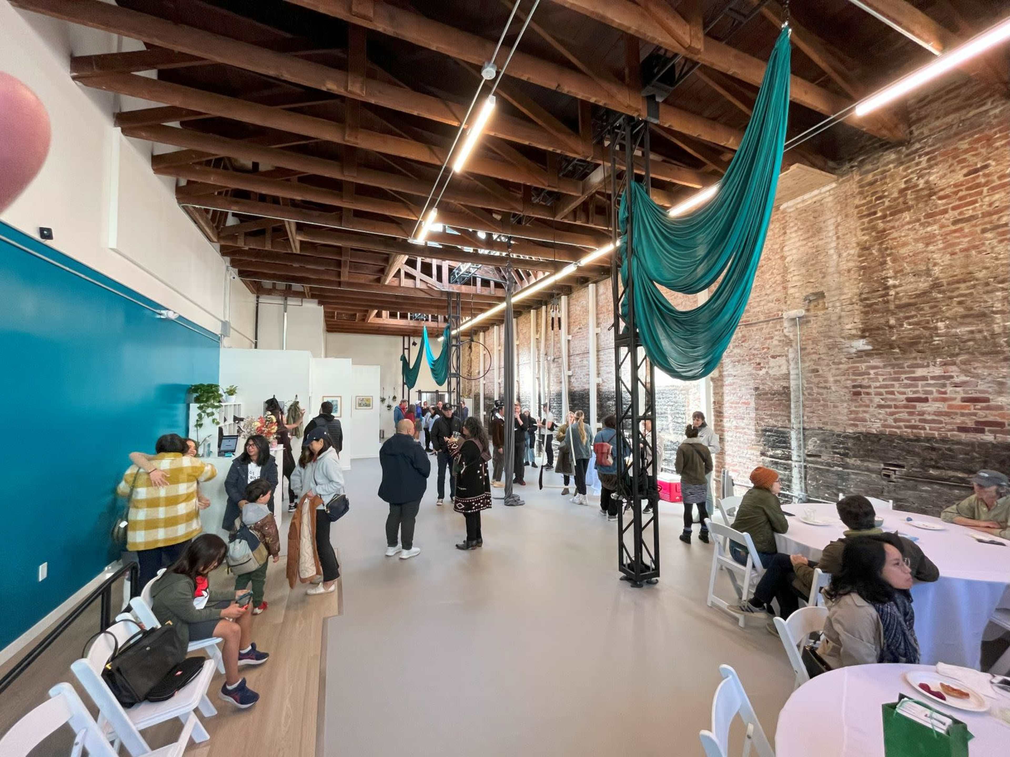 A spacious event space with exposed brick walls and wooden beams, filled with people mingling and seated at white folding tables.