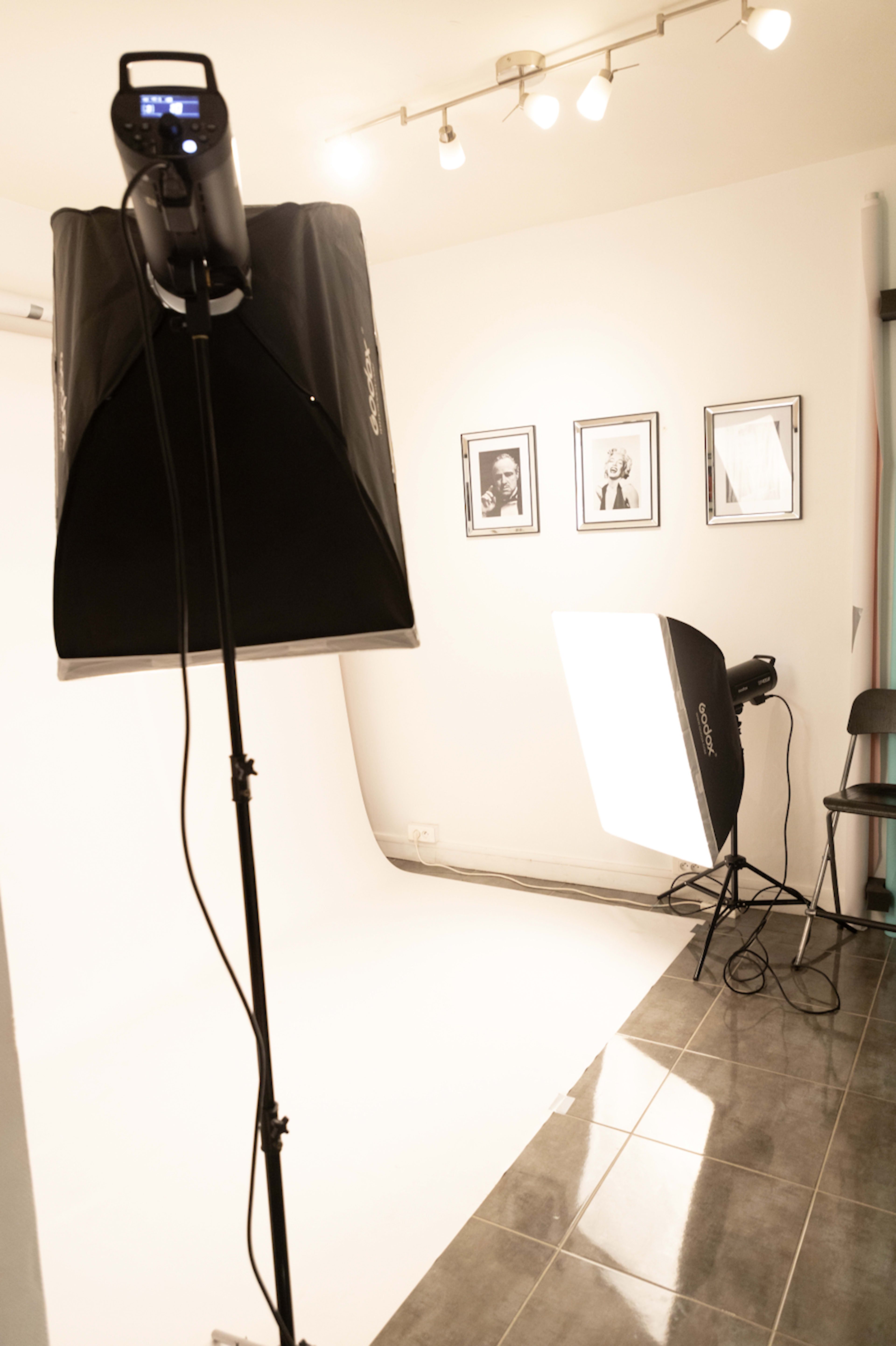 A photography studio setup featuring a softbox light and a backdrop on a tiled floor with framed photographs on the wall.