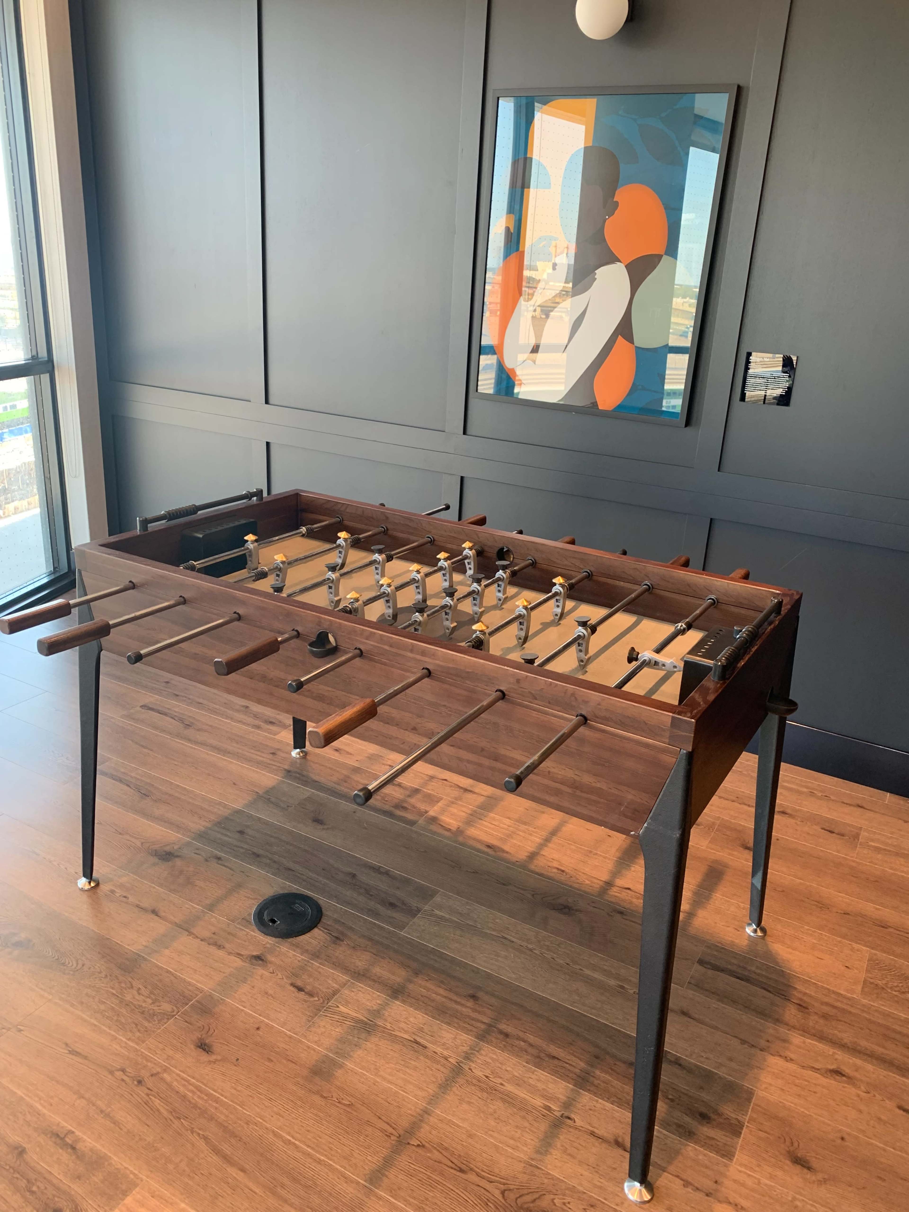 The image shows a foosball table placed in a room with dark panels and a colorful abstract artwork on the wall.
