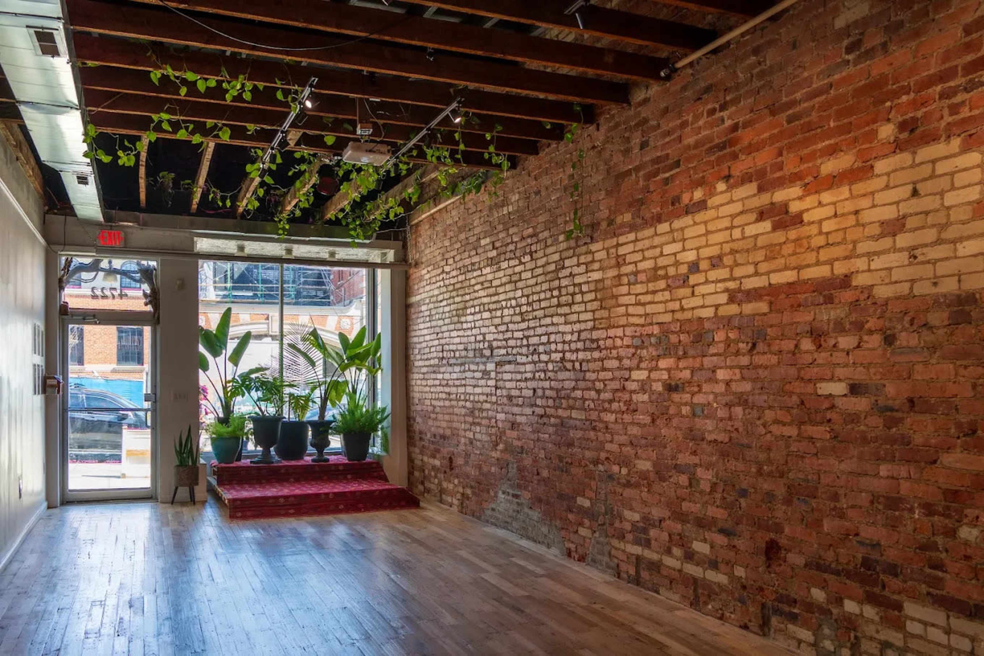 The image shows an interior space with exposed brick walls, wooden floors, and a doorway leading to a street, complemented by potted plants on a red carpet.