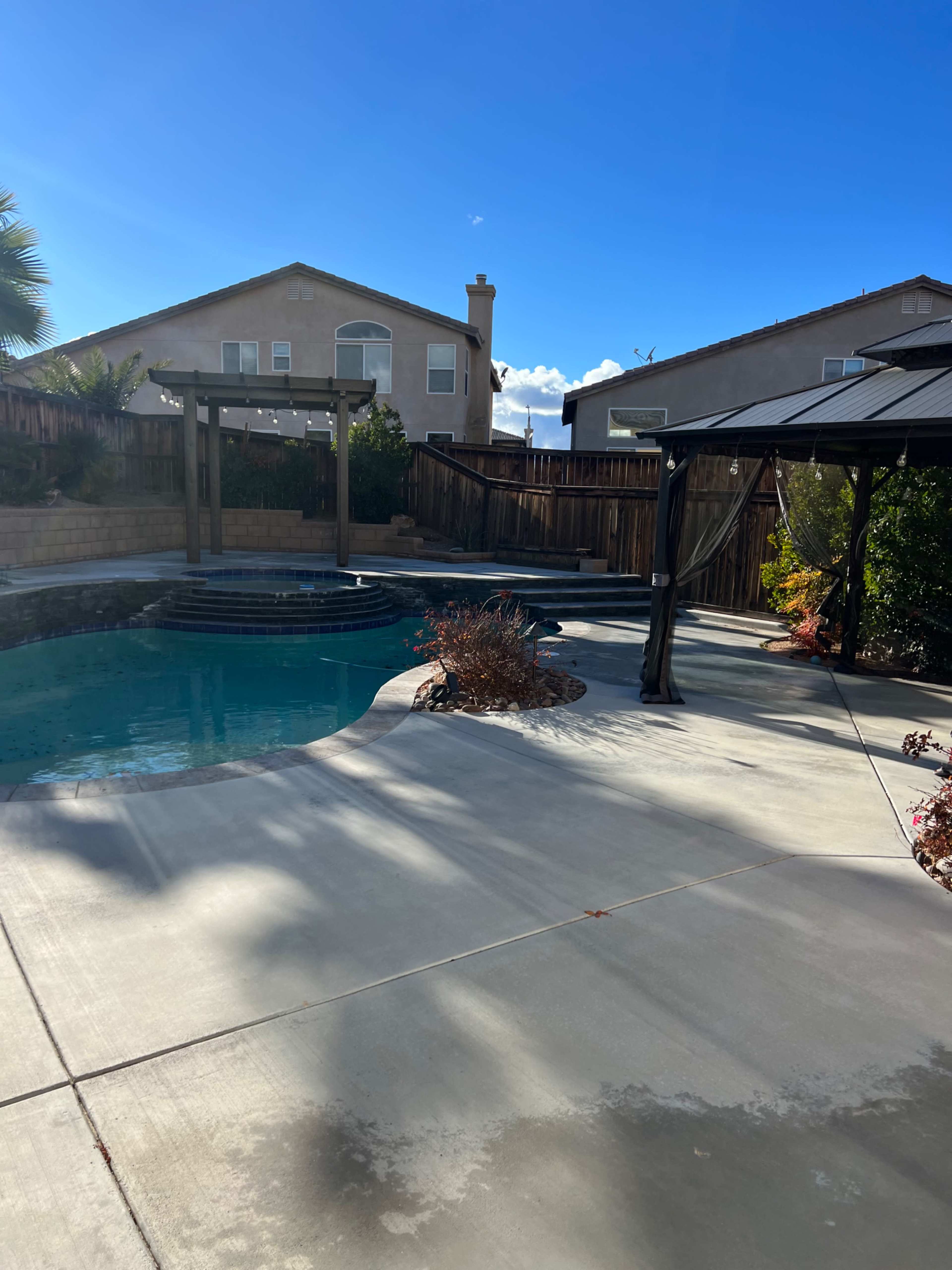 A backyard features a pool with a stone edge, surrounded by landscaping and a shaded gazebo under a clear blue sky.