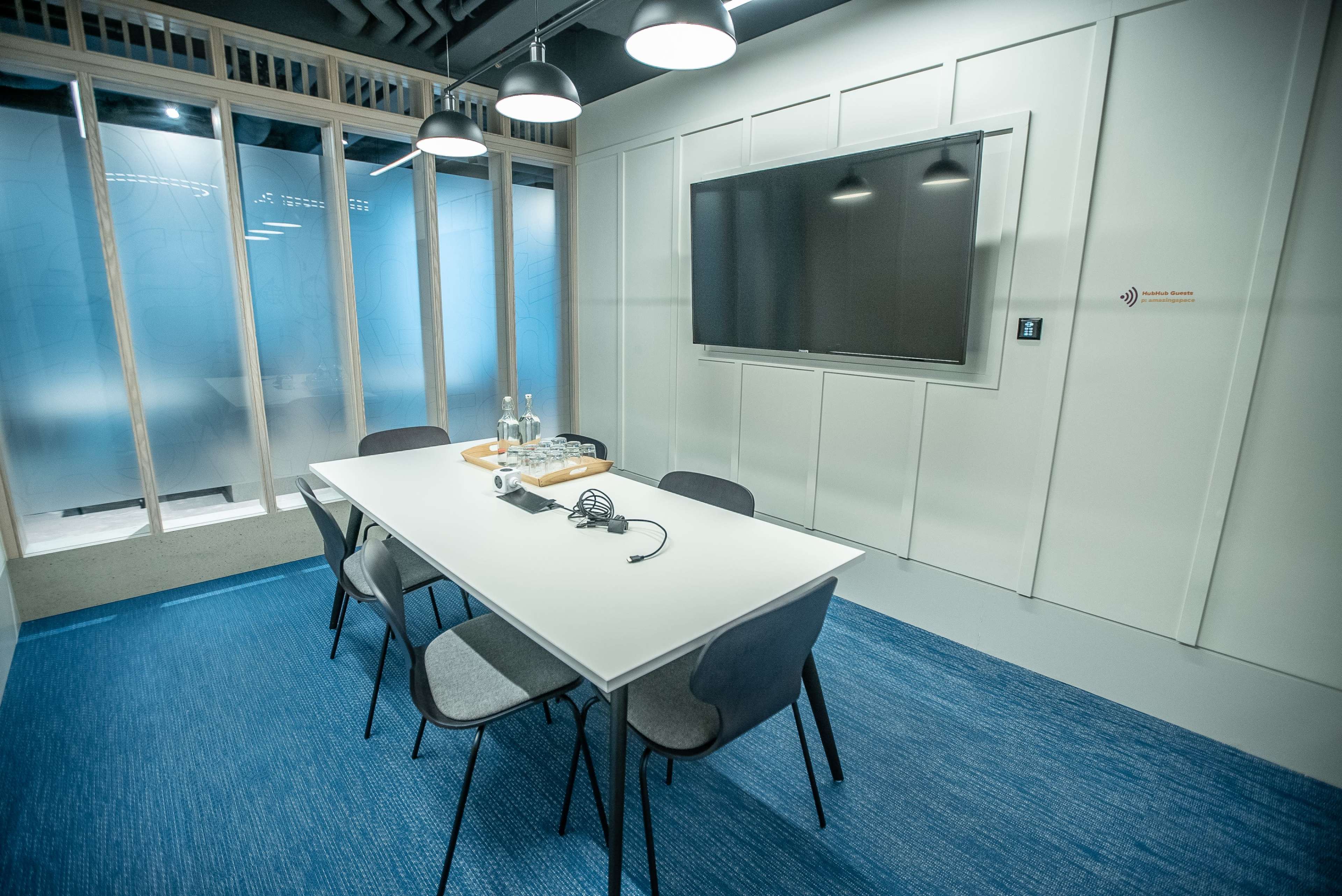 A modern meeting room features a long white table surrounded by gray chairs, with a large television mounted on the wall and a blue carpeted floor.