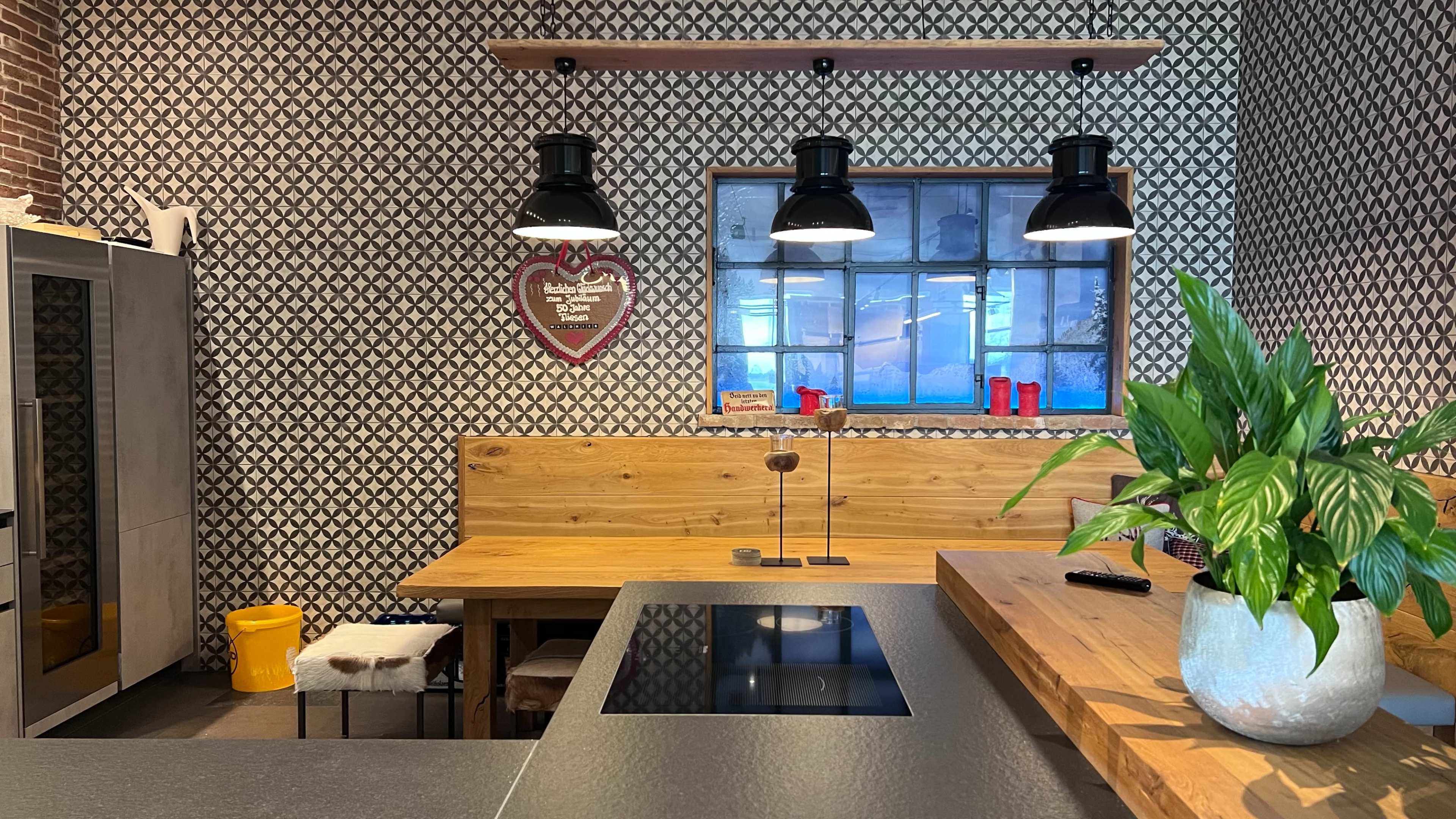 The image shows a modern kitchen with a patterned wall, a wooden dining table, and a large window above a countertop with black pendant lights.