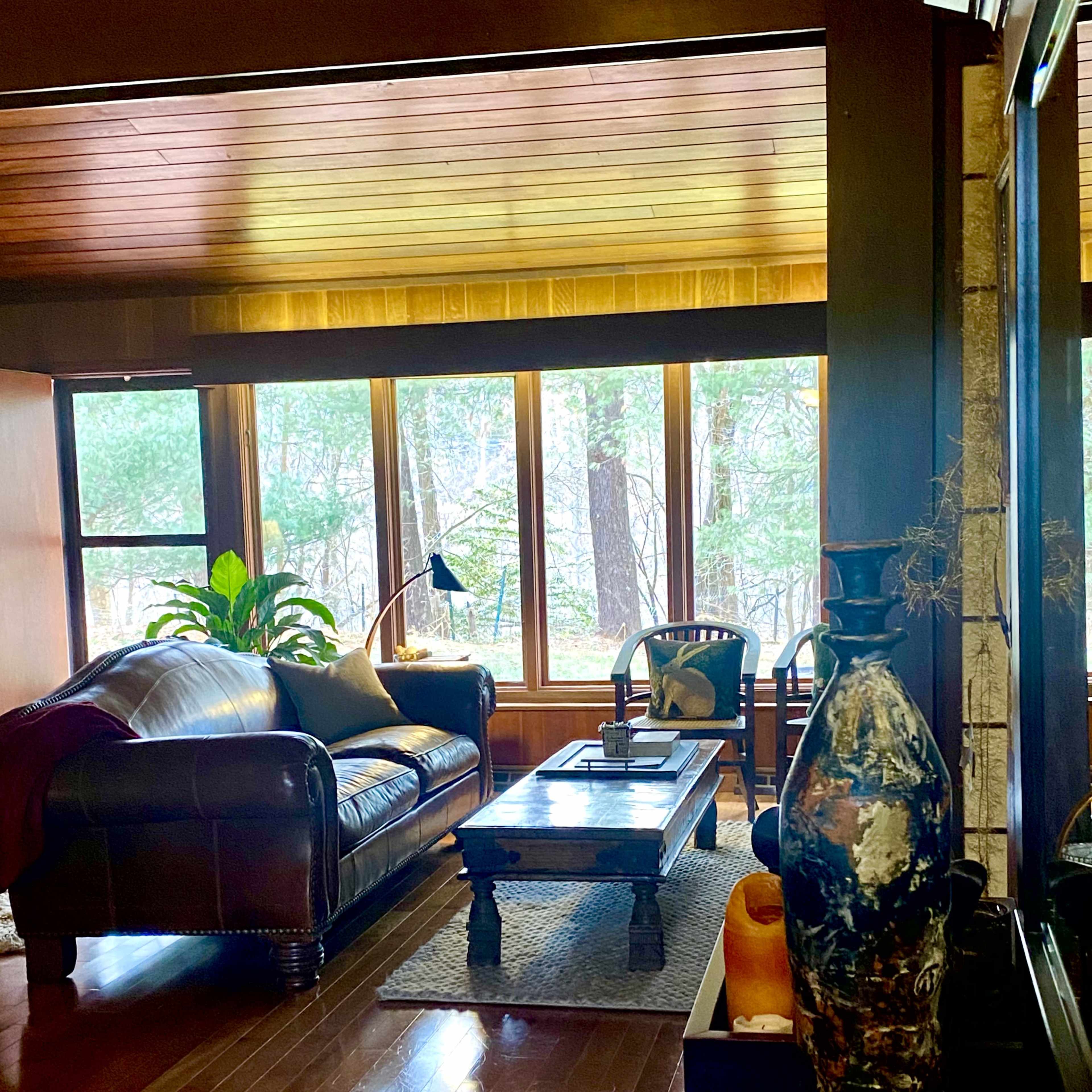 A cozy living room features a brown leather sofa, a wooden coffee table, and large windows overlooking a wooded area.