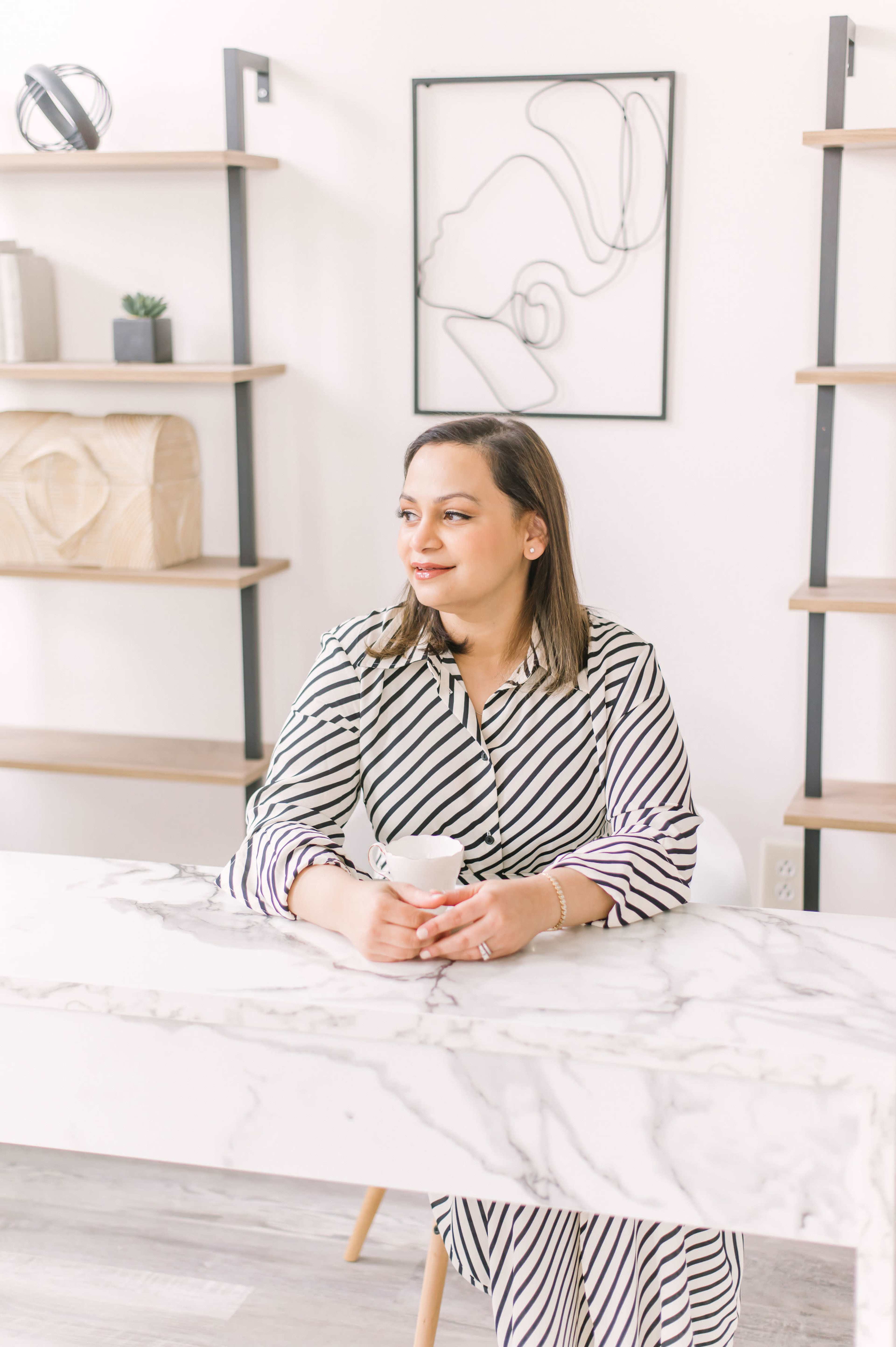 A woman seated at a marble table holds a cup while looking out, with modern shelving and a line drawing on the wall behind her.