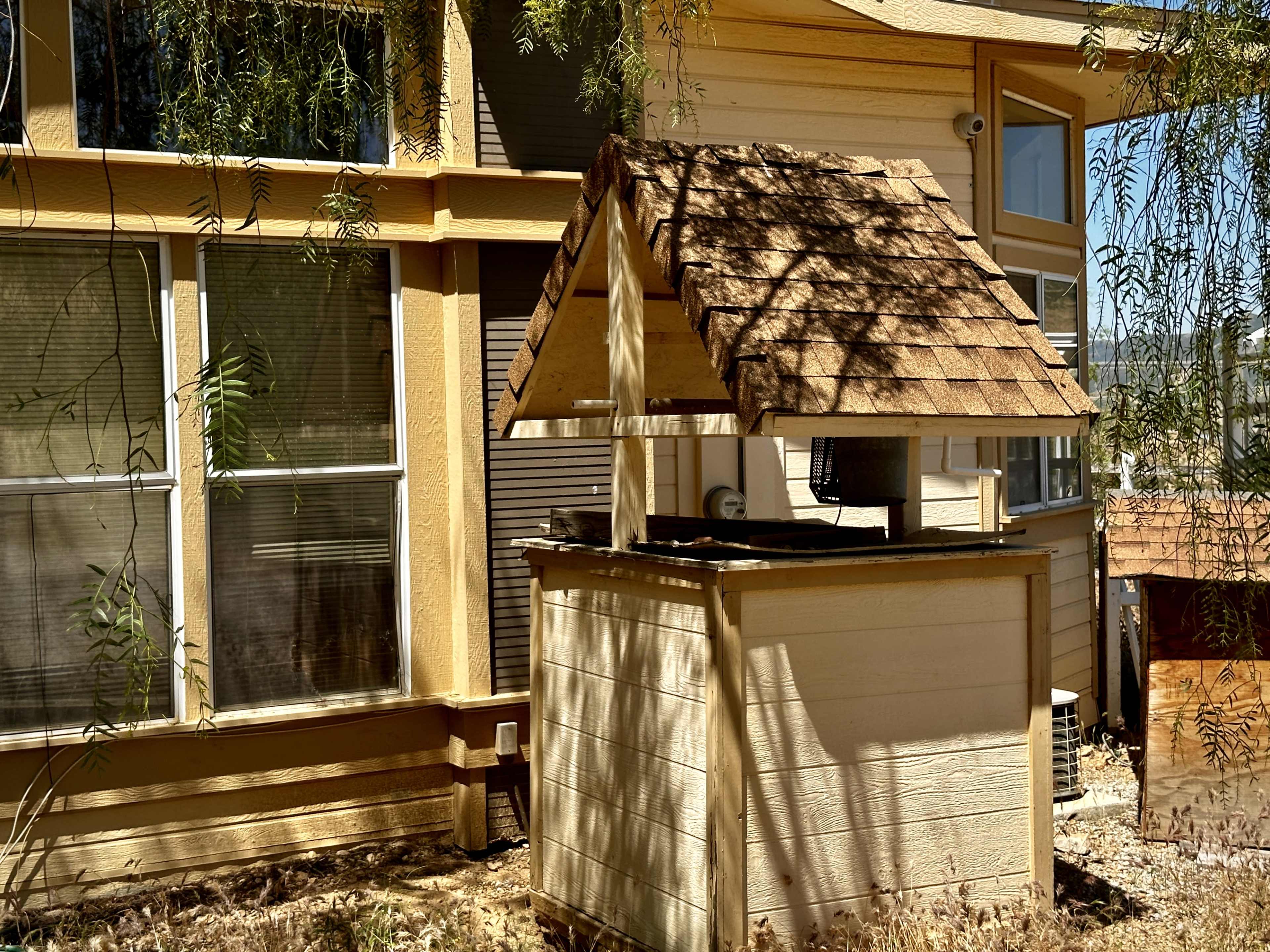 A wooden wishing well with a slanted roof, situated next to a house with large windows.