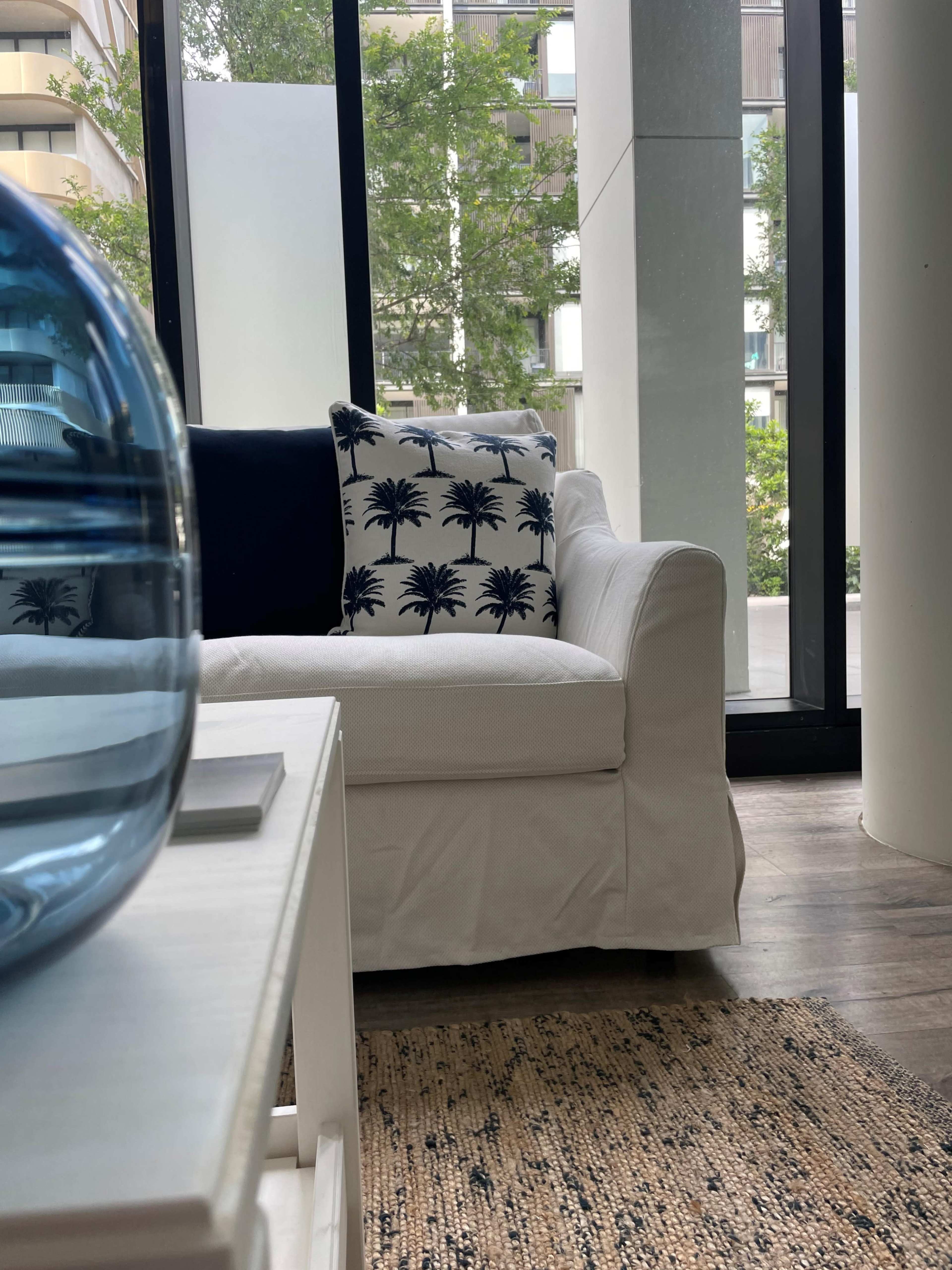 A white sofa with a palm tree-patterned pillow sits in a modern living space with large windows and a view of greenery outside.