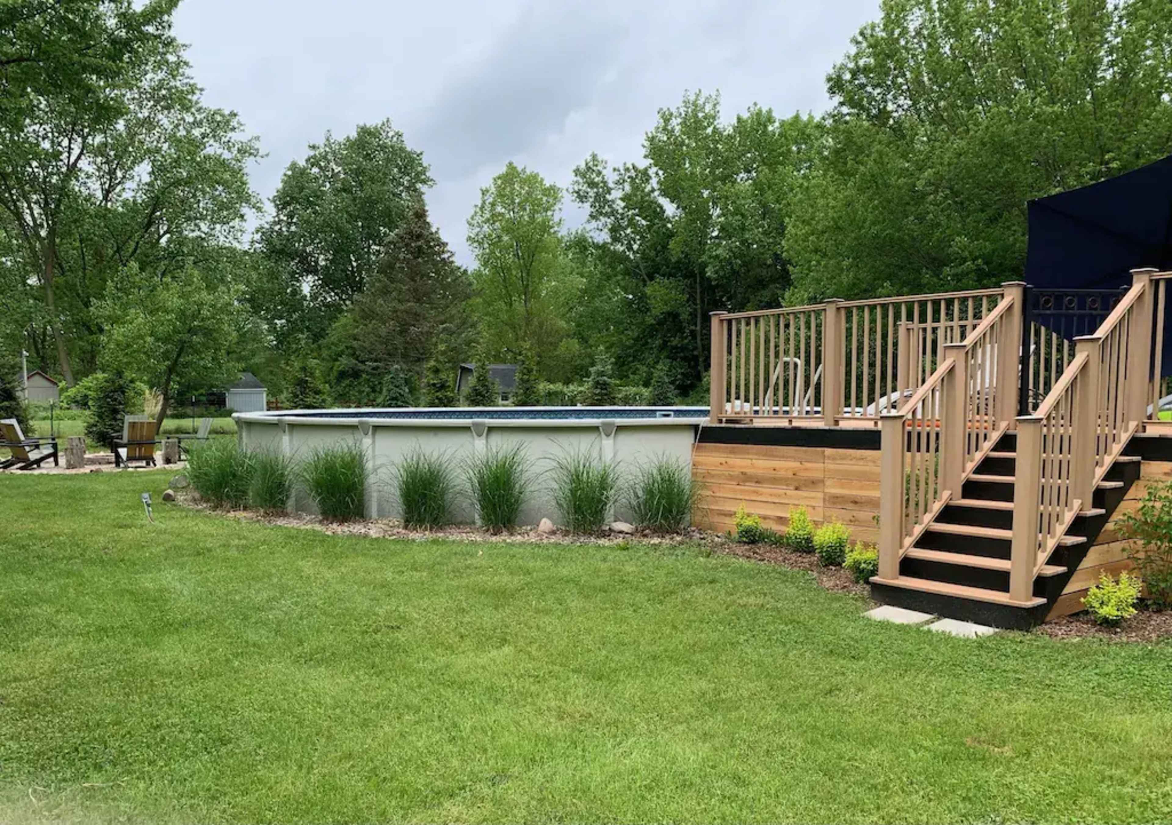A backyard with a round above-ground pool surrounded by grass, landscaping, and a wooden deck with stairs leading up to it.