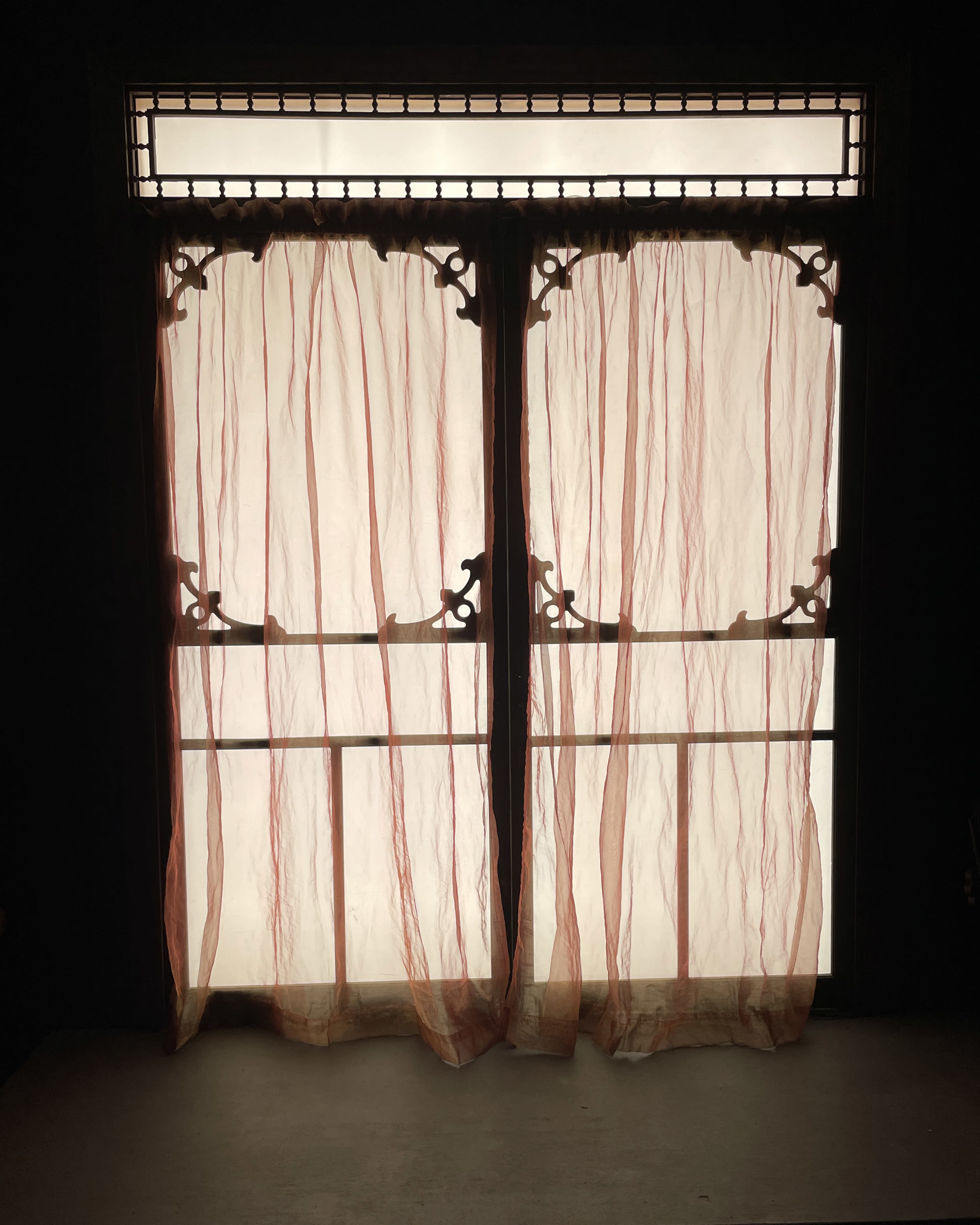 Sheer, lightly colored curtains hang in front of a window with an intricate frame, allowing soft light to filter through.