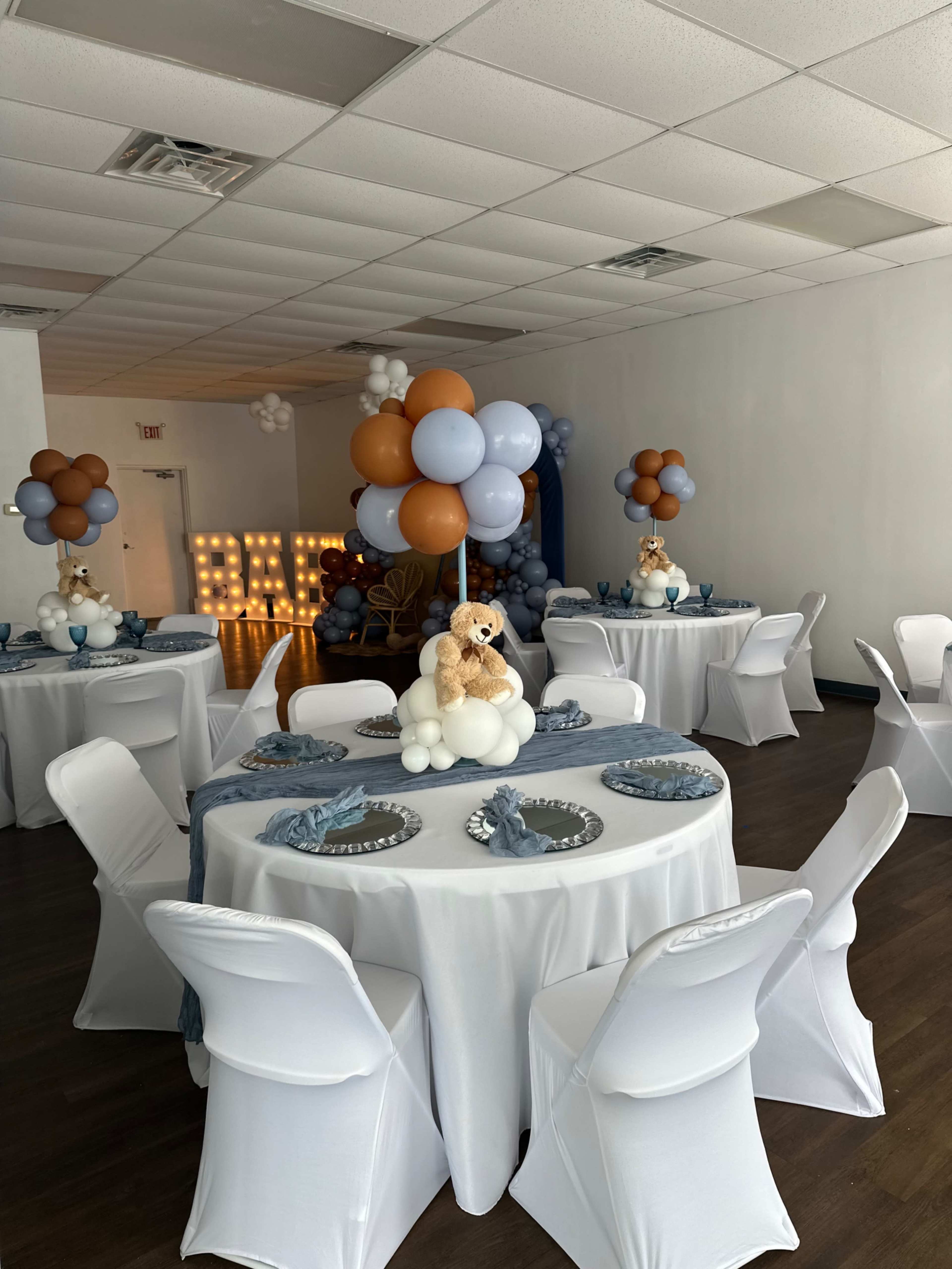 A decorated event space features round tables with white tablecloths, blue and brown balloon arrangements, and teddy bear centerpieces.