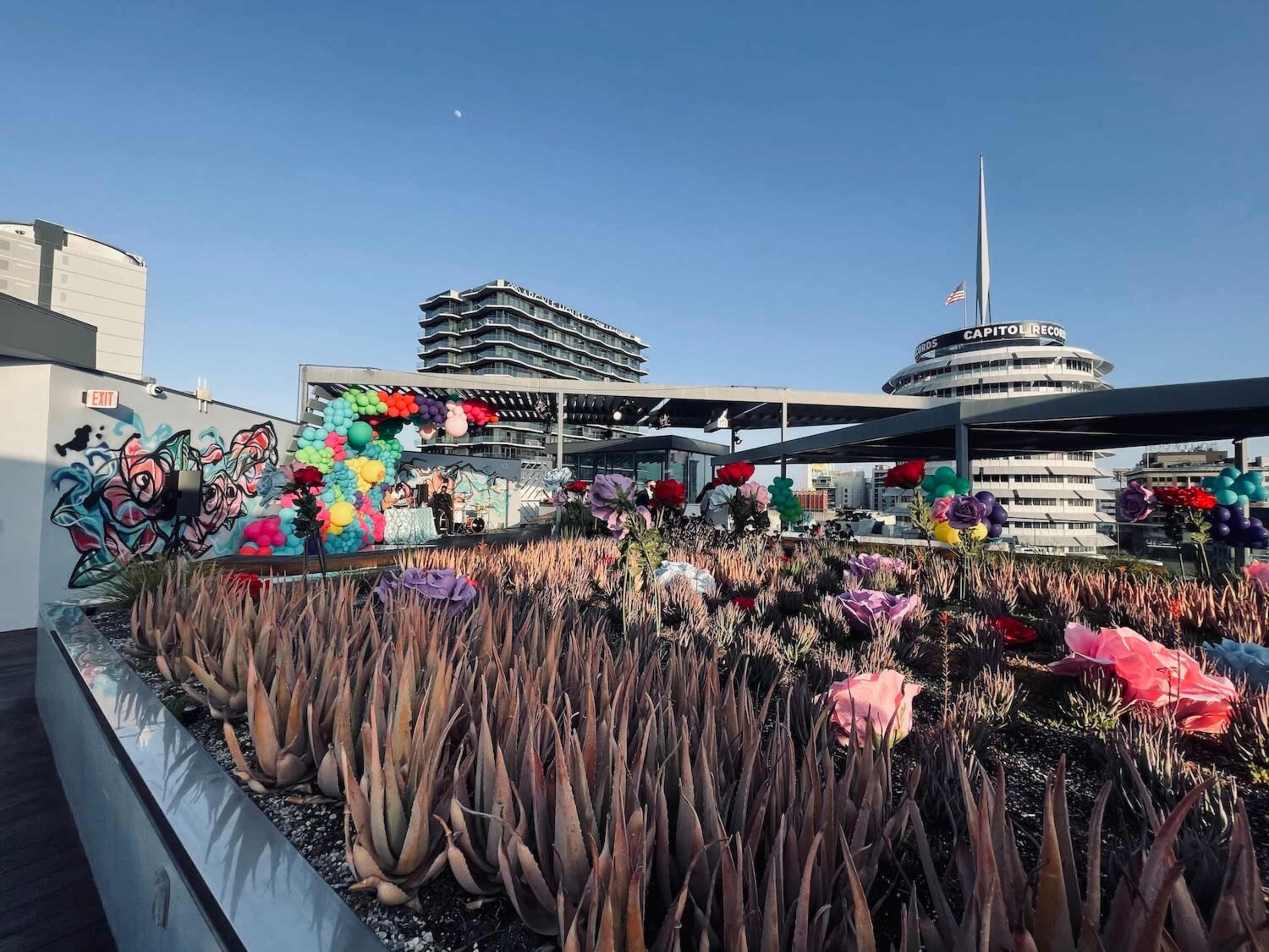 The image shows a rooftop garden adorned with various colorful flowers and balloons, set against a backdrop of a modern building and the Capitol Records tower.