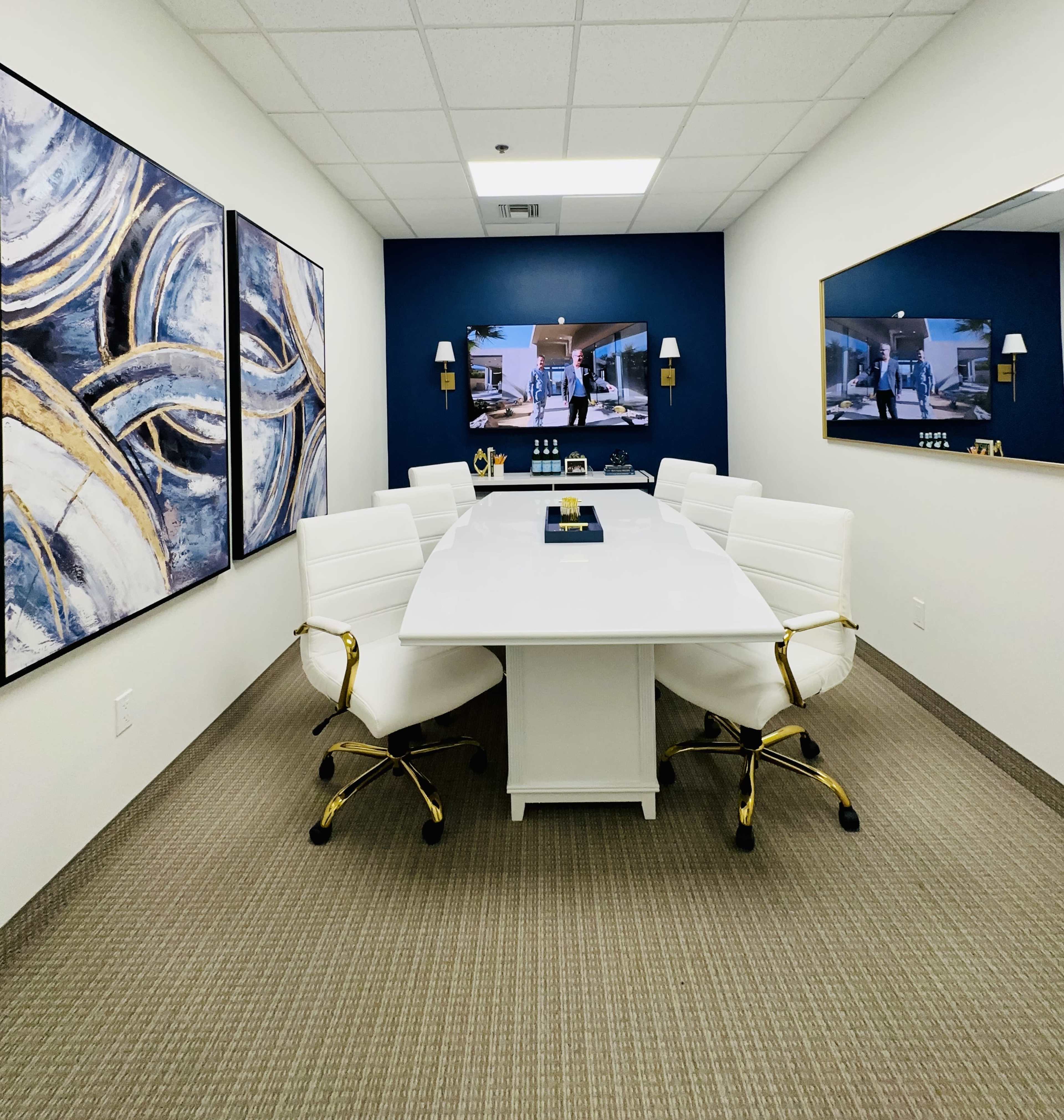 A modern conference room features a large white table surrounded by white chairs, with abstract artworks on the walls and two monitors displaying visuals.