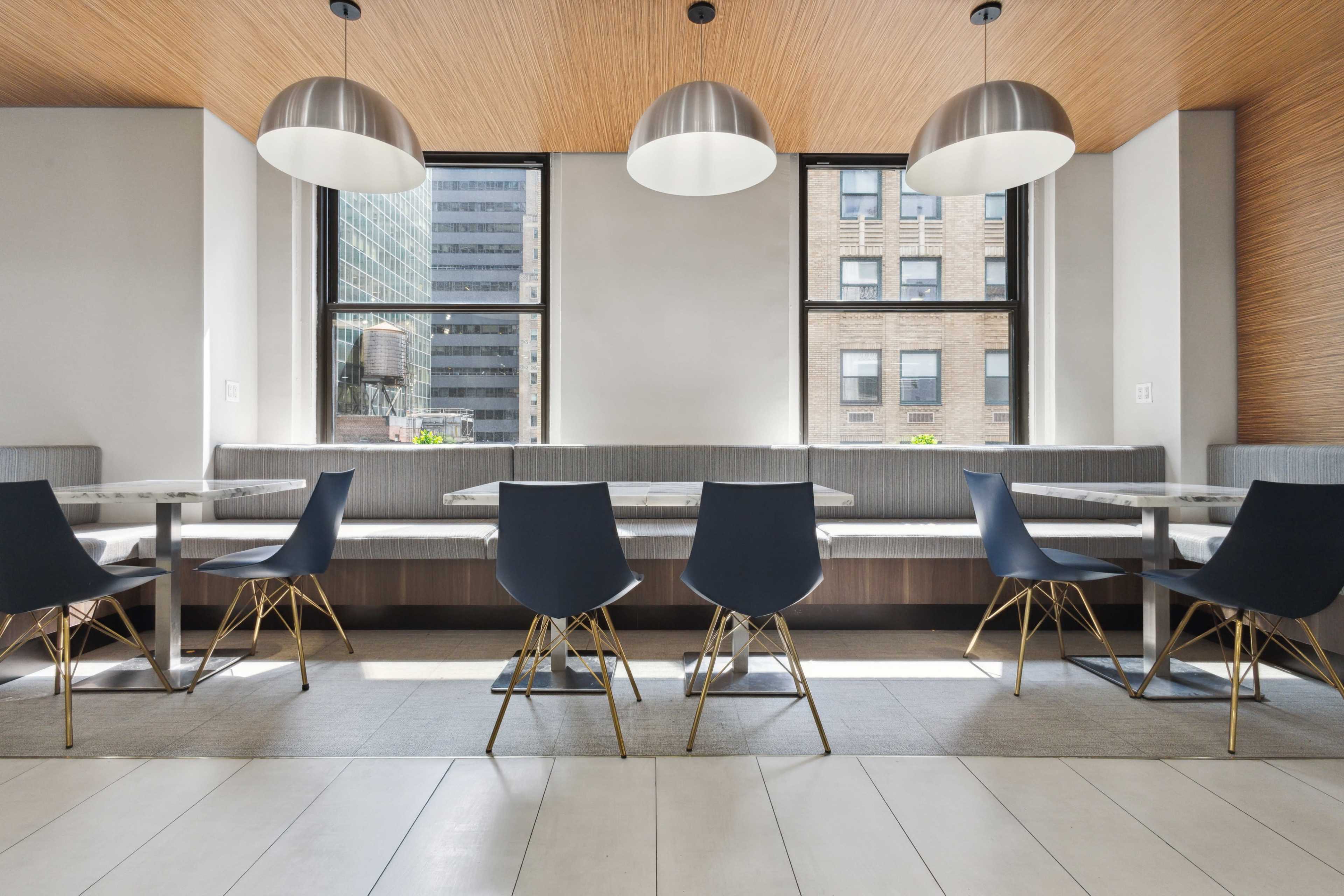 The image depicts a modern dining area featuring a long table with several chairs beneath large pendant lights and windows overlooking a cityscape.