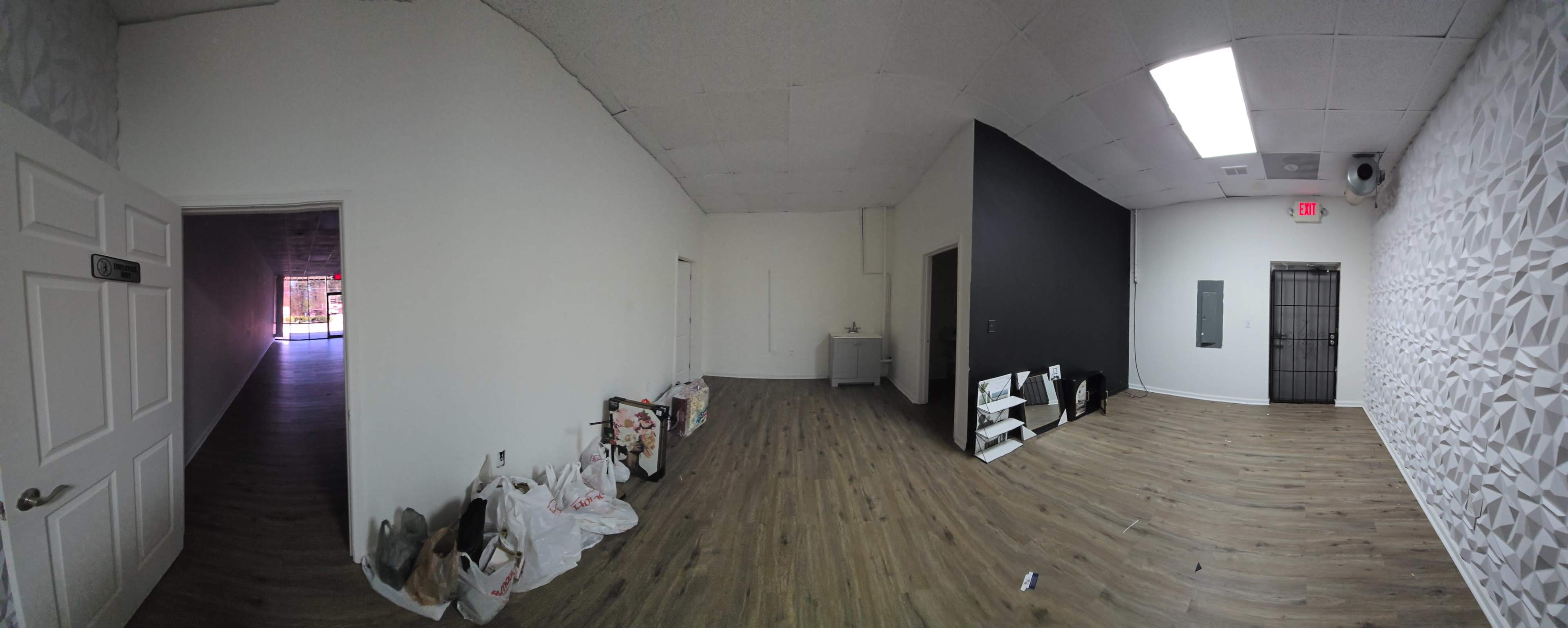 The image shows a spacious, empty room with wooden flooring and a black accent wall, featuring discarded items on the floor and an adjoining hallway leading to another area.
