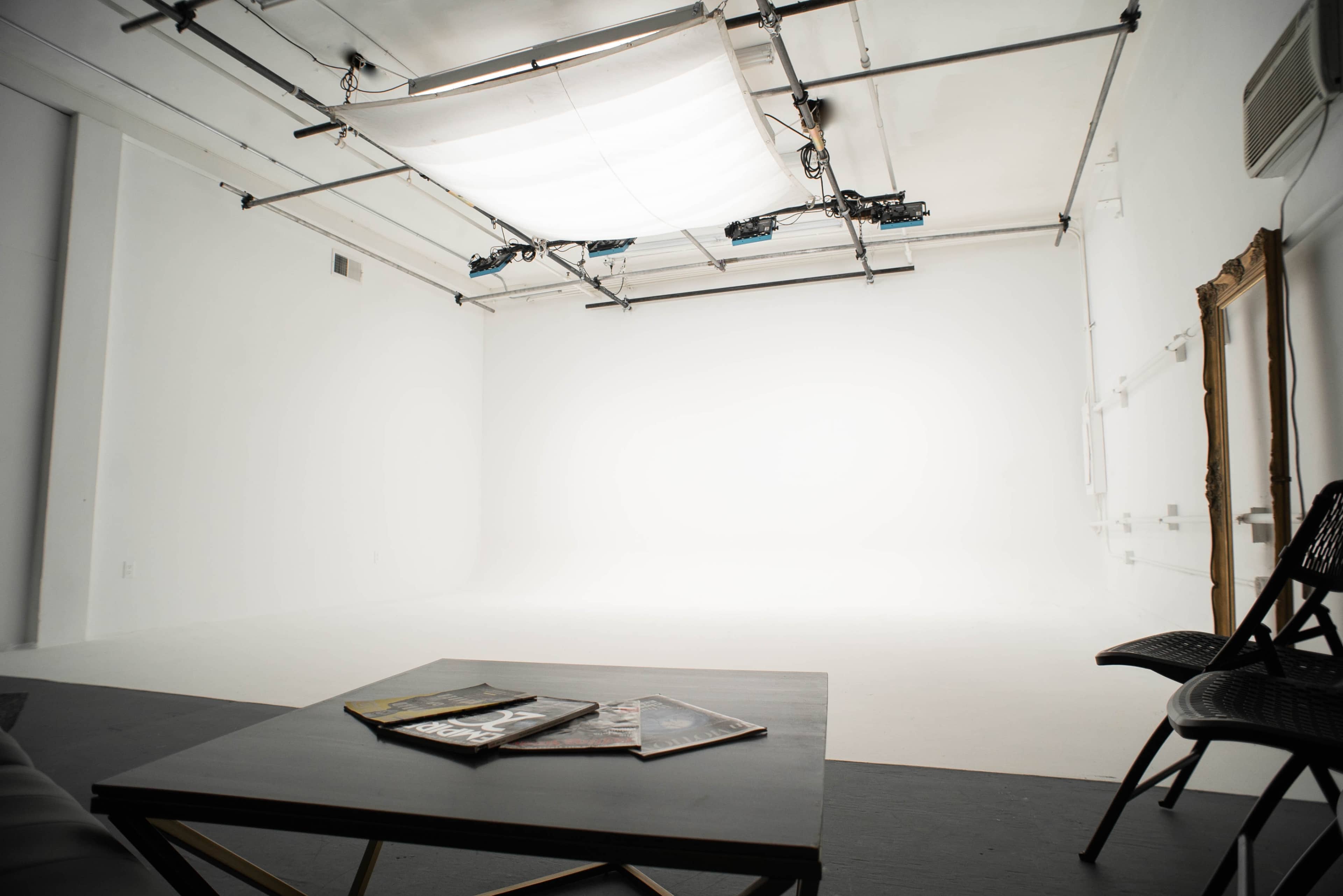 A minimalist studio space with a large white backdrop, a low table with magazines, and a chair positioned against the wall.