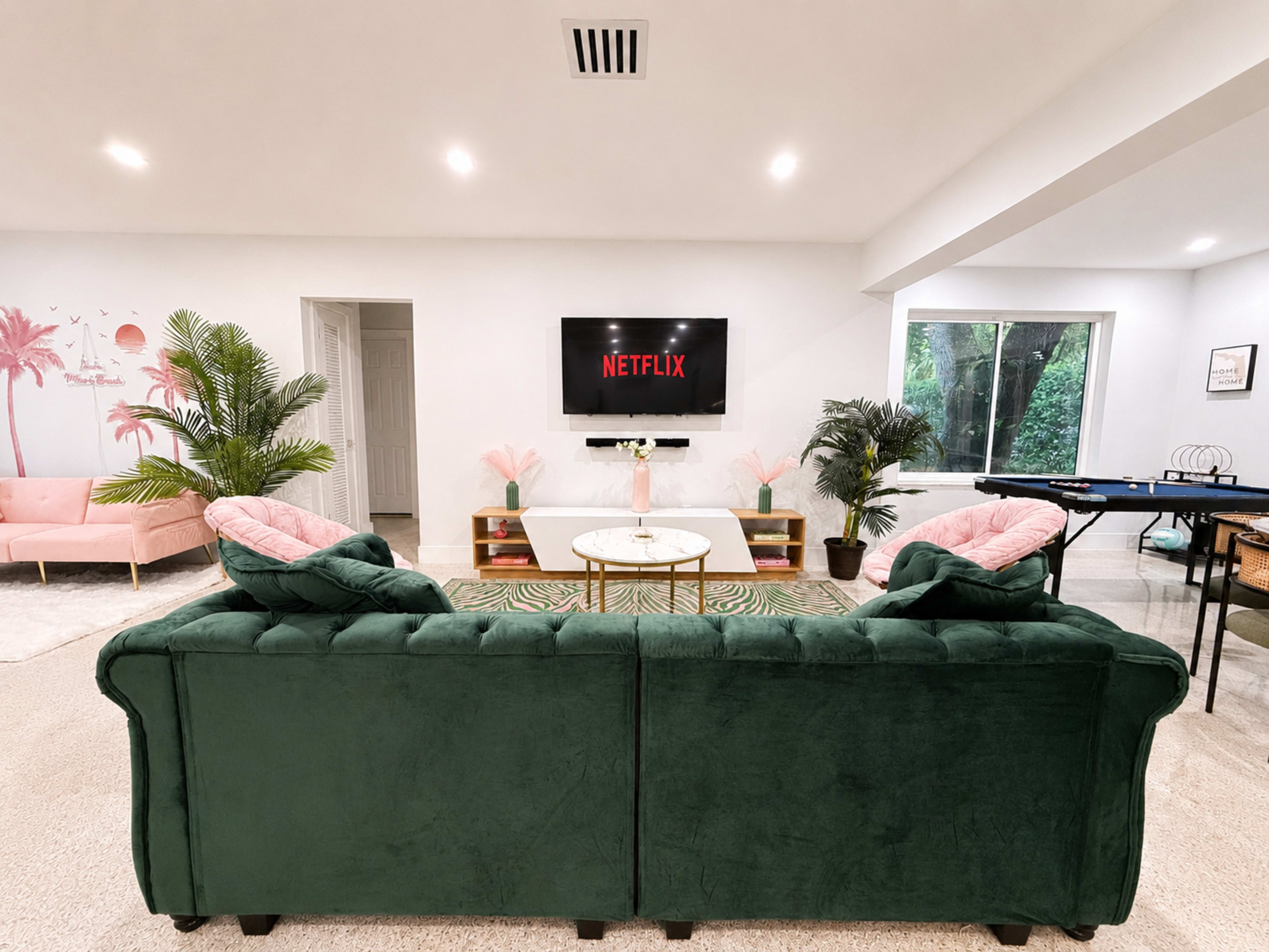 A cozy living room features a green couch facing a wall-mounted TV displaying the Netflix logo, with pink accents and tropical plants throughout the space.