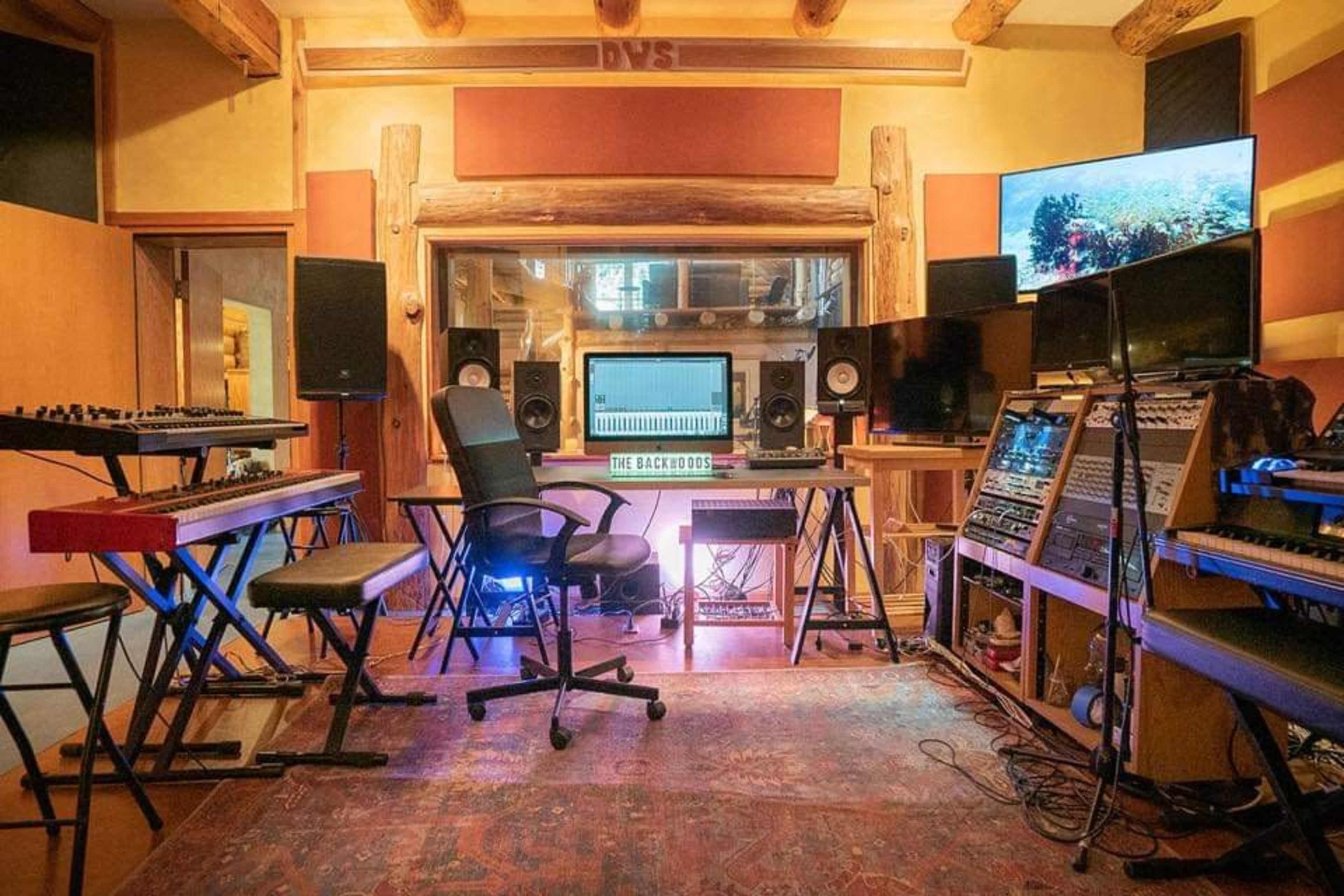 A well-equipped music studio featuring various keyboards, audio monitors, a central computer workstation, and sound equipment arranged around a patterned area rug.