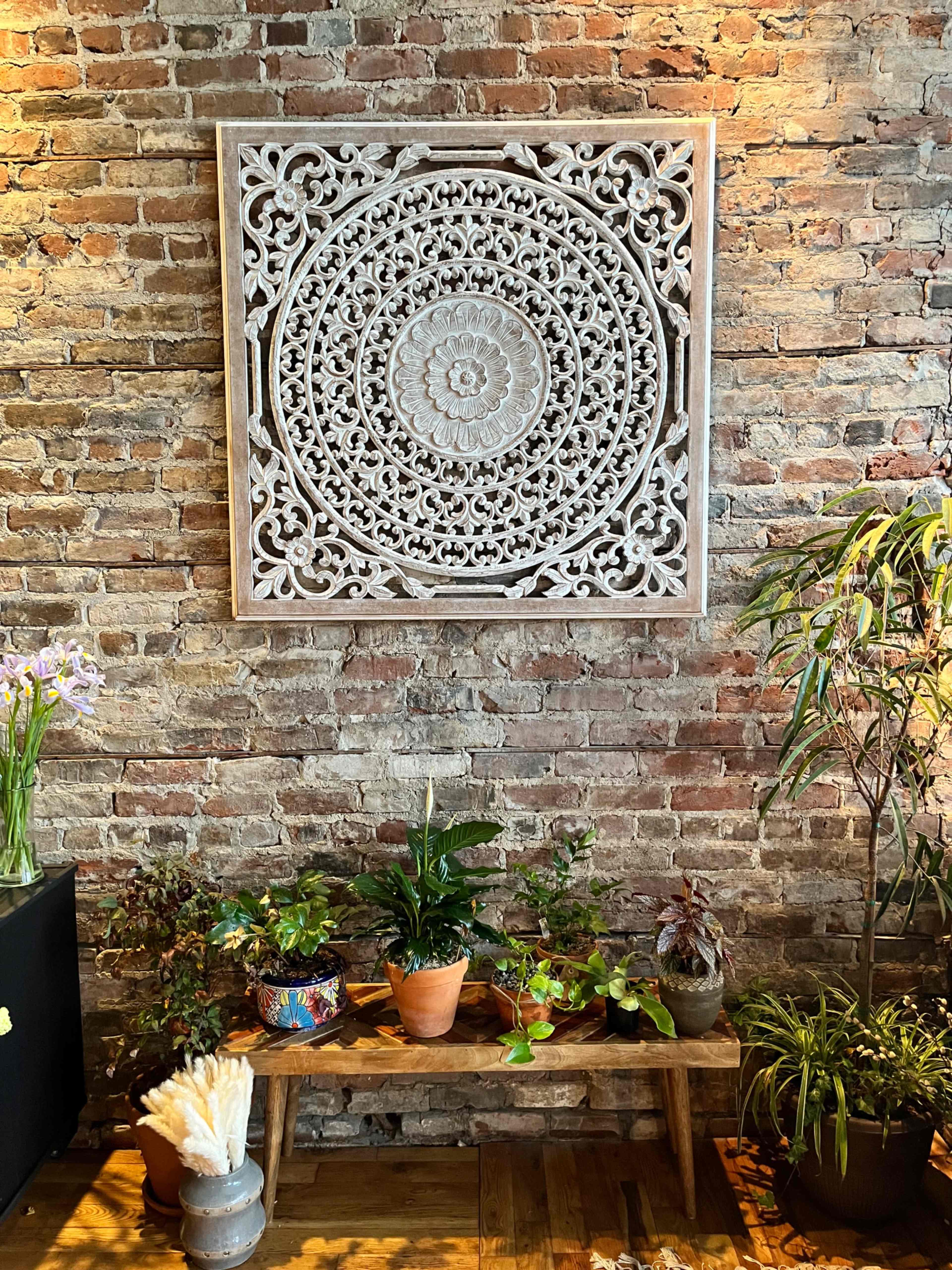 A carved wooden wall decoration is mounted on a brick wall above a wooden bench with several potted plants.