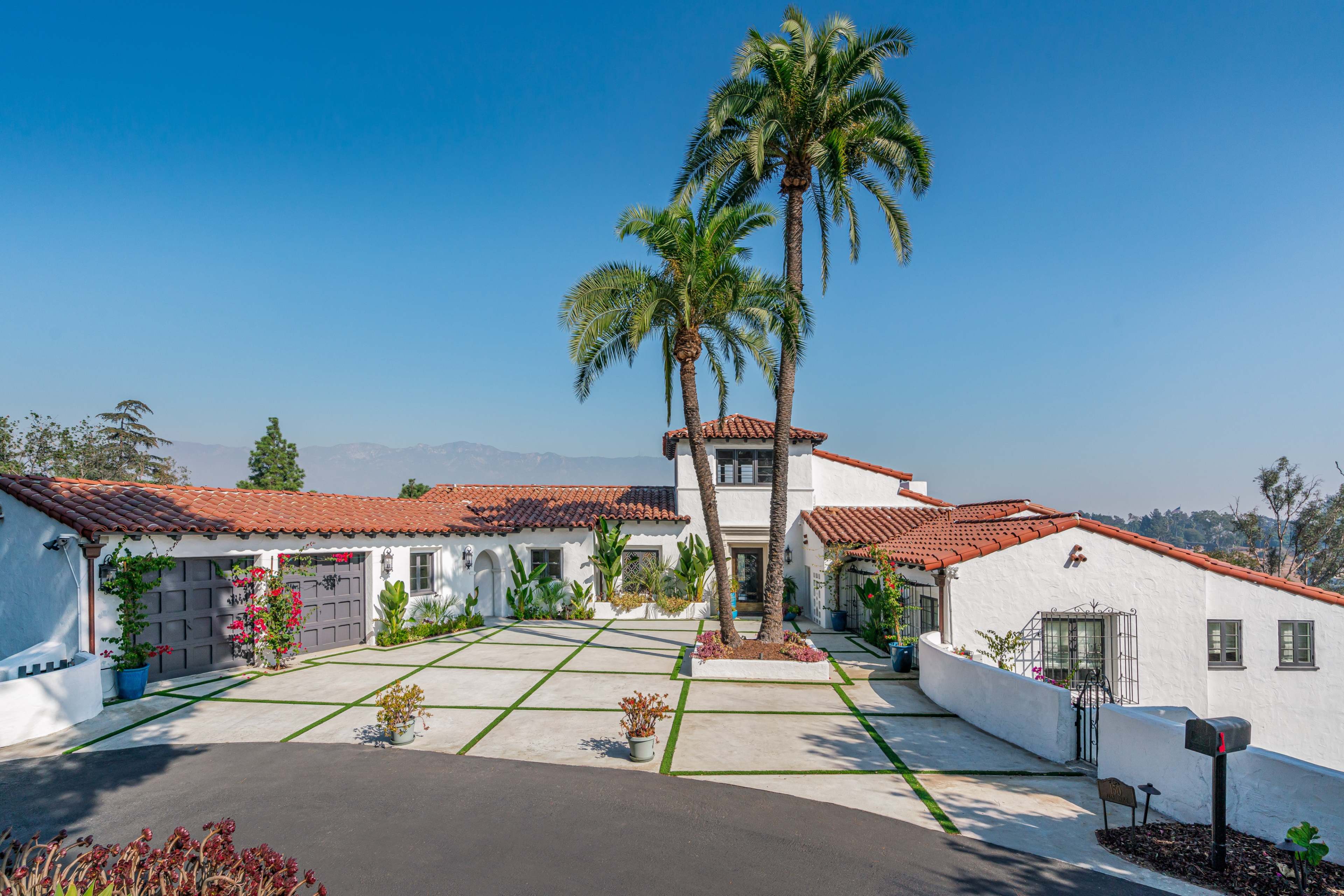 A spacious, modern Mediterranean-style house with a red tile roof, surrounded by palm trees and landscaped gardens.