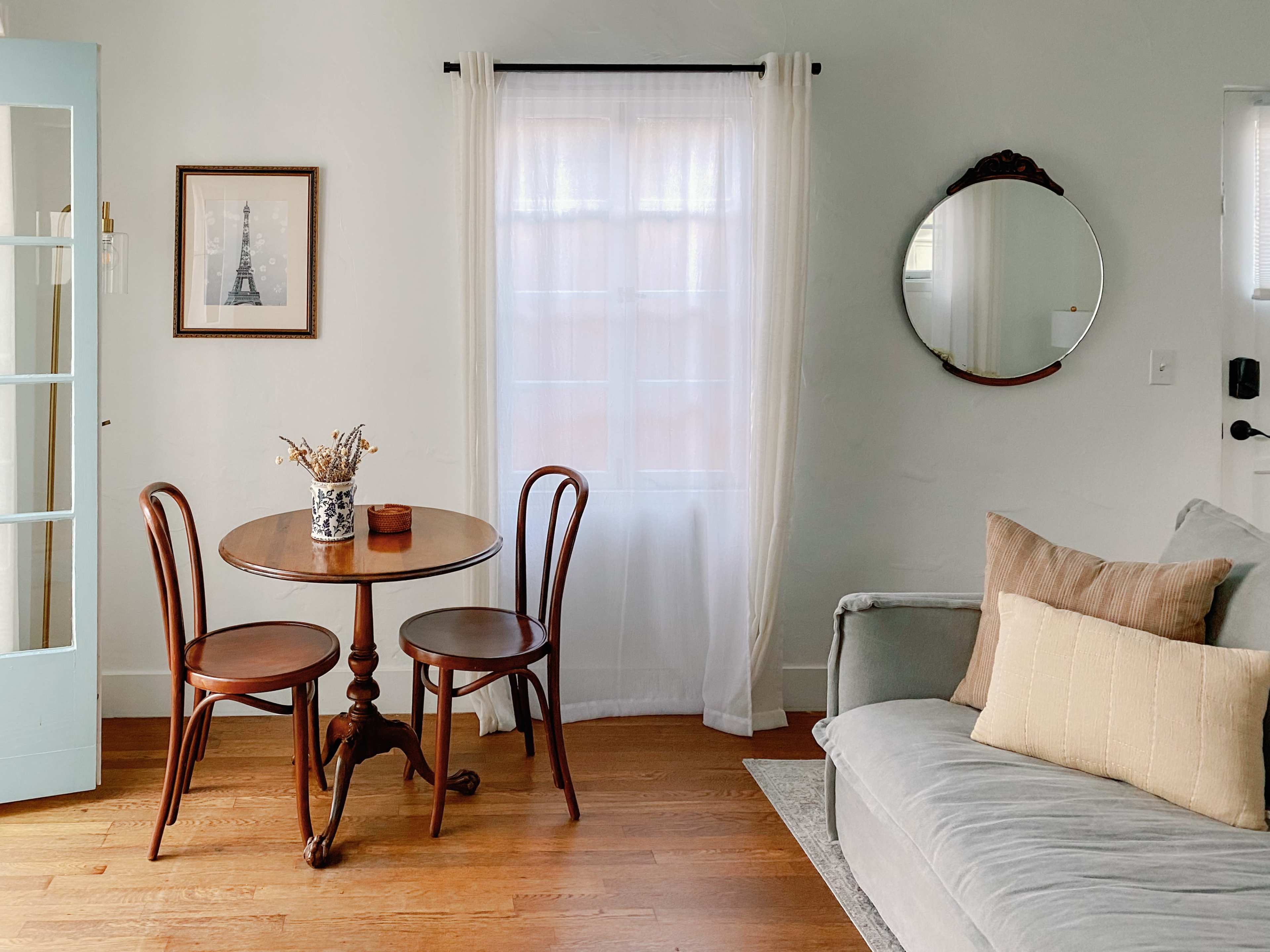 A small dining table with two chairs is positioned next to a window in a well-lit room featuring a gray sofa and a round mirror on the wall.
