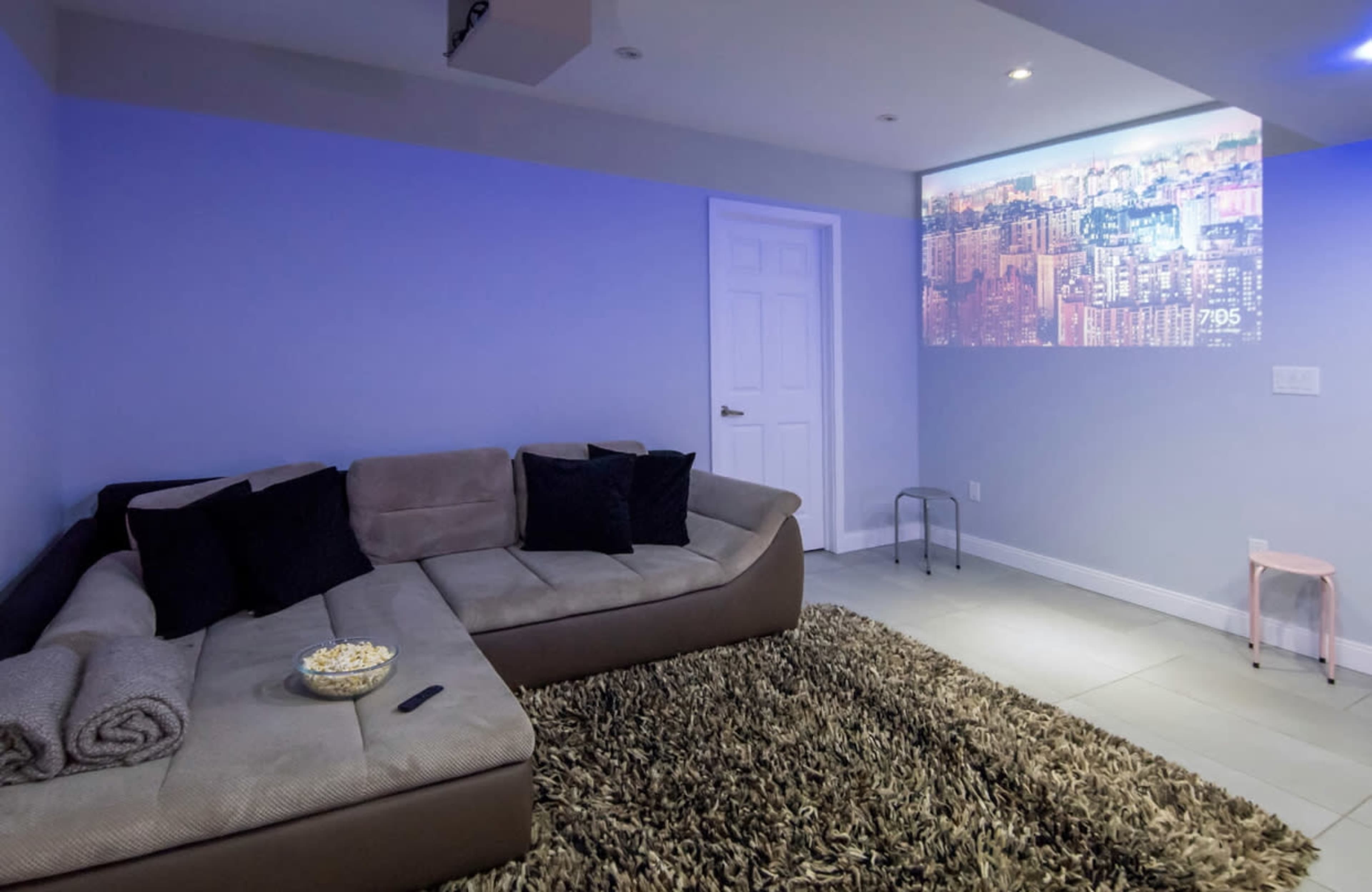 A cozy living room features a sectional sofa with black pillows, a projector displaying a cityscape on the wall, and a small table beside it with a bowl of popcorn.