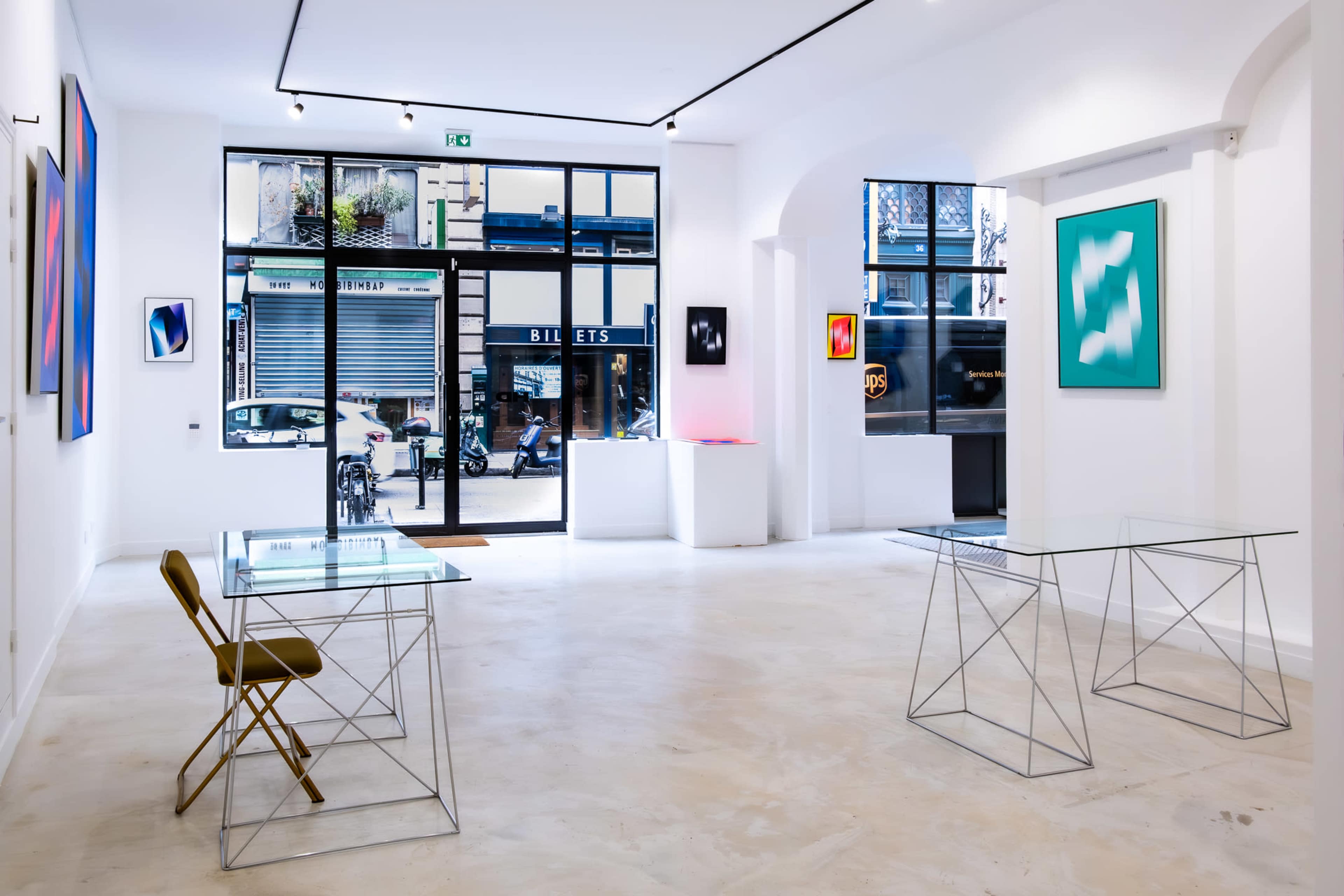 A contemporary art gallery featuring two glass-topped tables with metal bases, a couple of framed artworks on the walls, and large windows allowing natural light to enter.