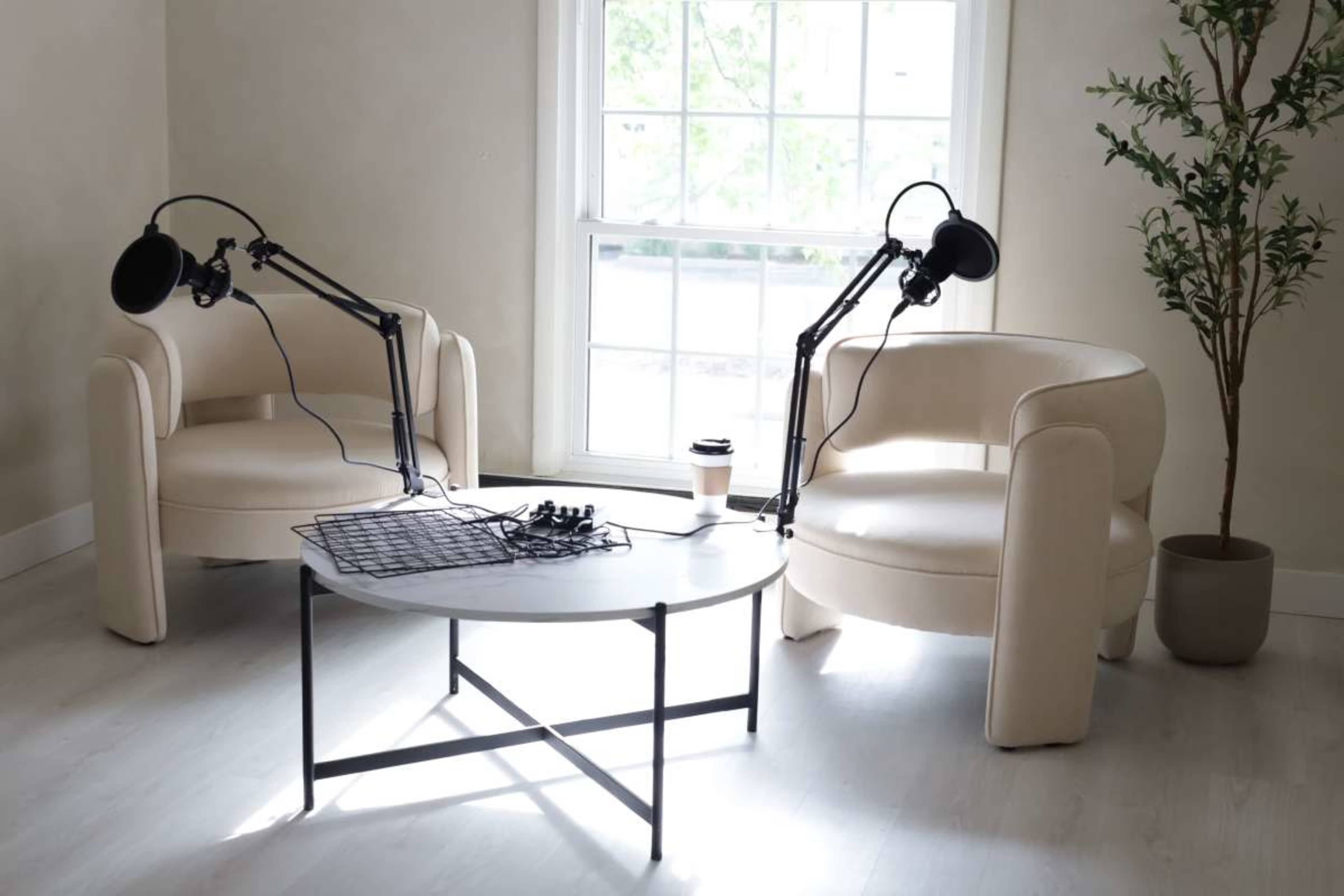 Two light-colored armchairs with adjustable microphone stands positioned beside a round coffee table in a well-lit room.